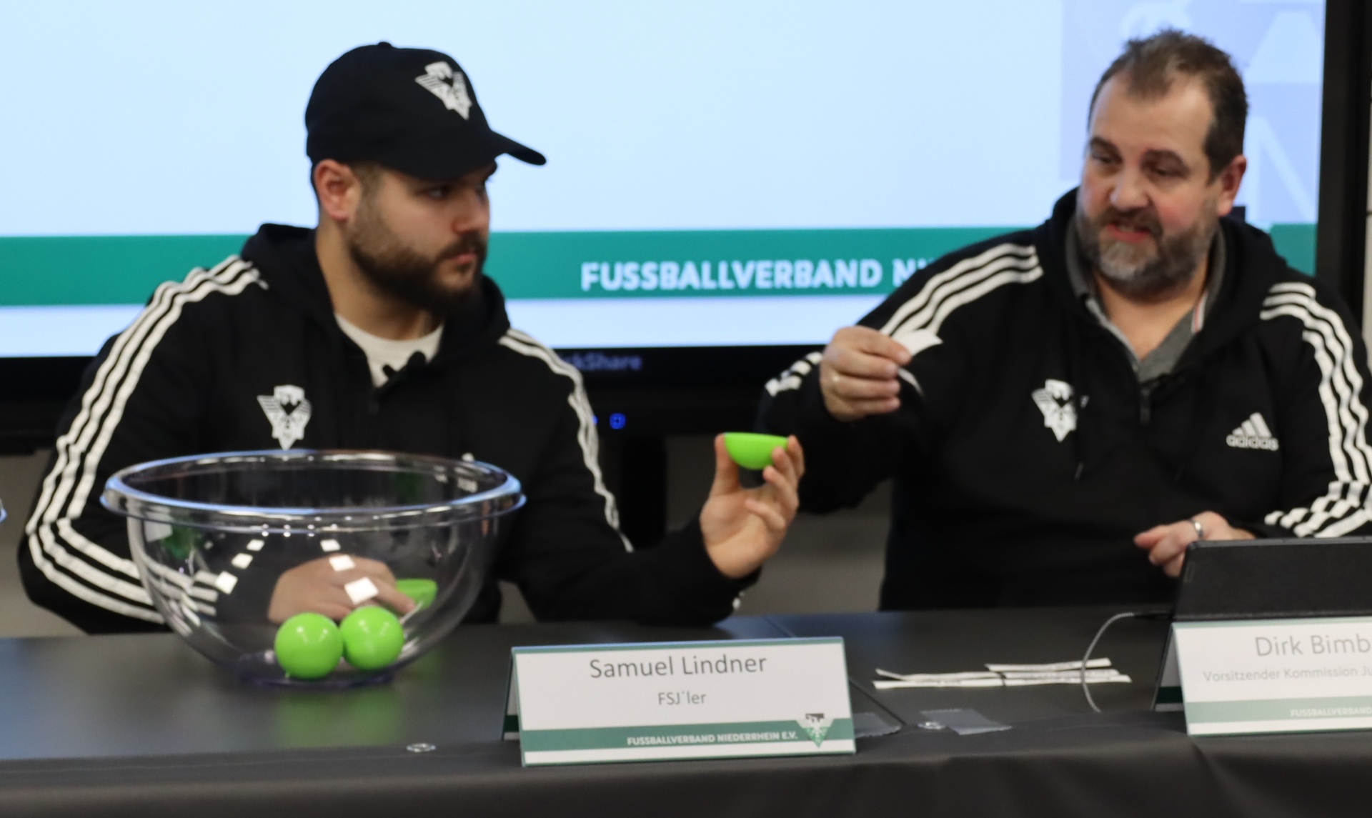 [{"clang_id":1,"value":"Zwei Männer sitzen an einem Tisch. Links trägt einer Kappe und Jacke, hält grüne Bälle. Rechts sitzt ein Mann mit Bart, ebenfalls in Jacke, mit einem Ball. Vor ihnen Namensschilder."}]