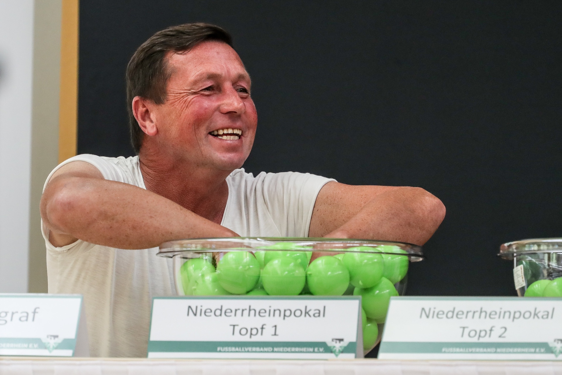[{"clang_id":1,"value":"Mann zieht an zwei durchsichtigen Behältern mit grünen Bällen. Davor stehen Schilder mit der Aufschrift \"Niederrheinpokal Topf 1\" und \"Topf 2\"."}]