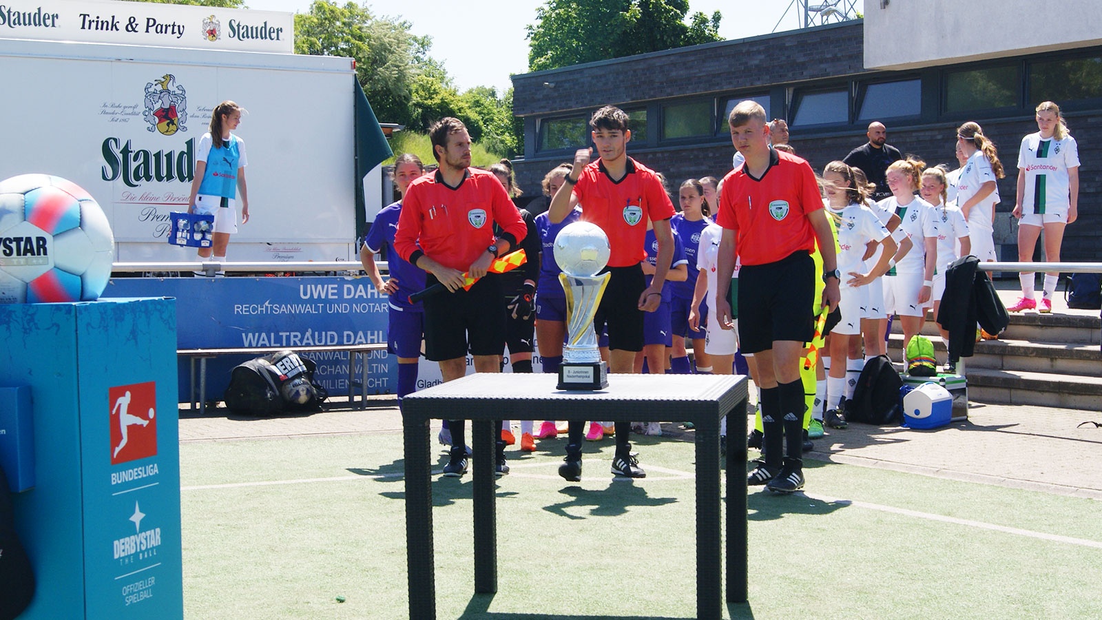 [{"clang_id":1,"value":"Fußballmannschaft mit Trainer vor weißem Gebäude. Eine Person hält einen Pokal. Im Hintergrund Werbebanner. Rasenfläche im Vordergrund."}]