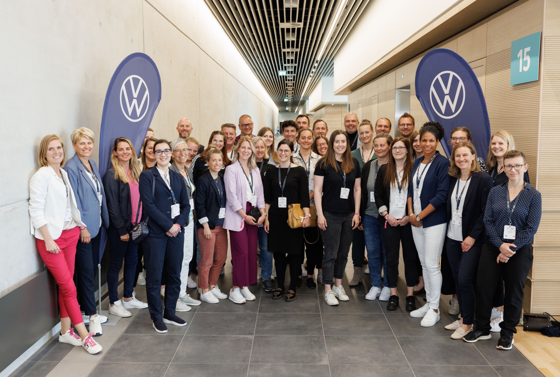 [{"clang_id":1,"value":"Gruppe von etwa 30 Personen steht in einem hellen, modernen Gang mit Volkswagen-Logos. Viele tragen Namensschilder und sind leger gekleidet."}]