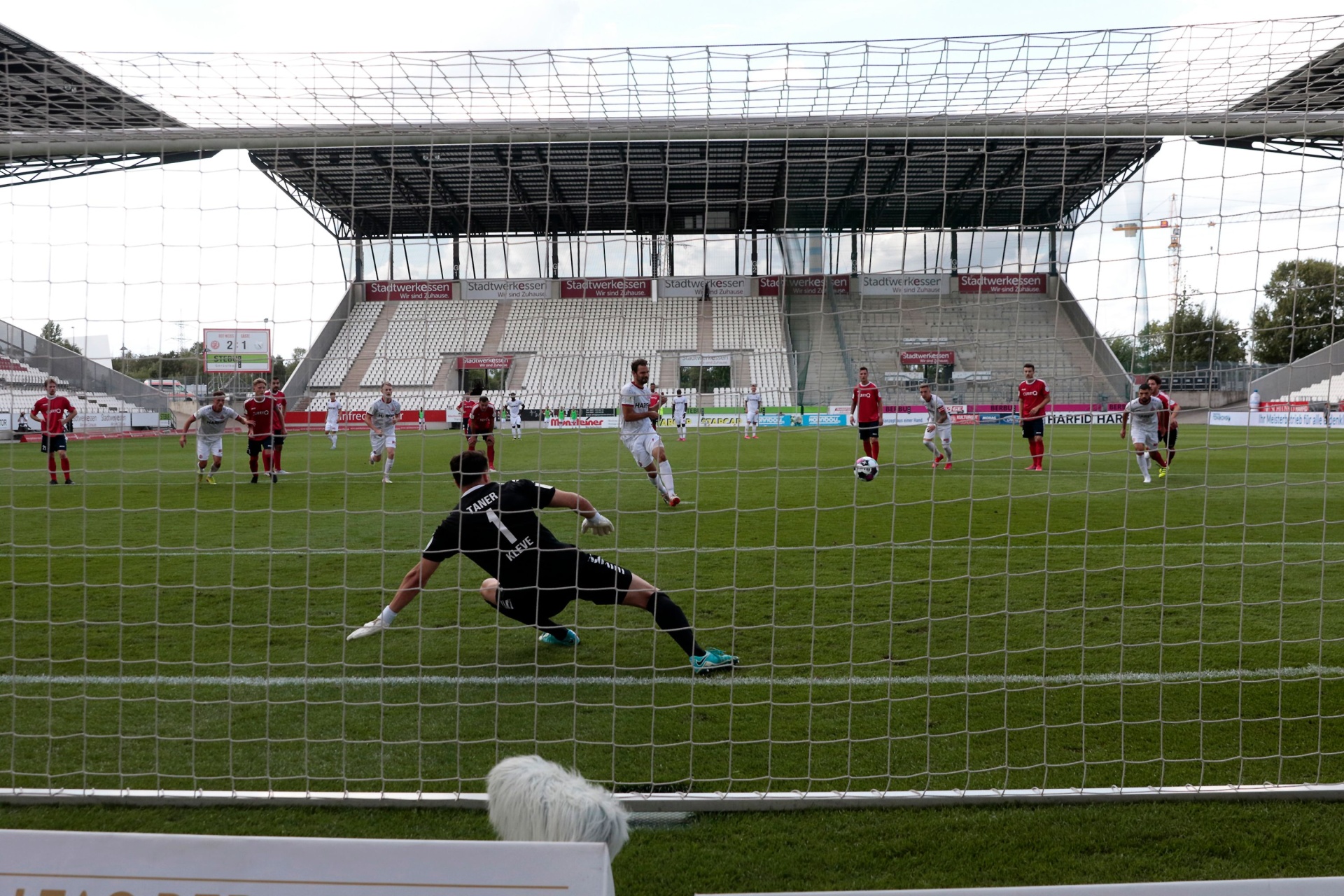[{"clang_id":1,"value":"Fußballspiel im Stadion. Spieler in roten und schwarzen Trikots auf grünem Rasen. Torwart springt, um Ball zu halten. Zuschauerreihen im Hintergrund."}]