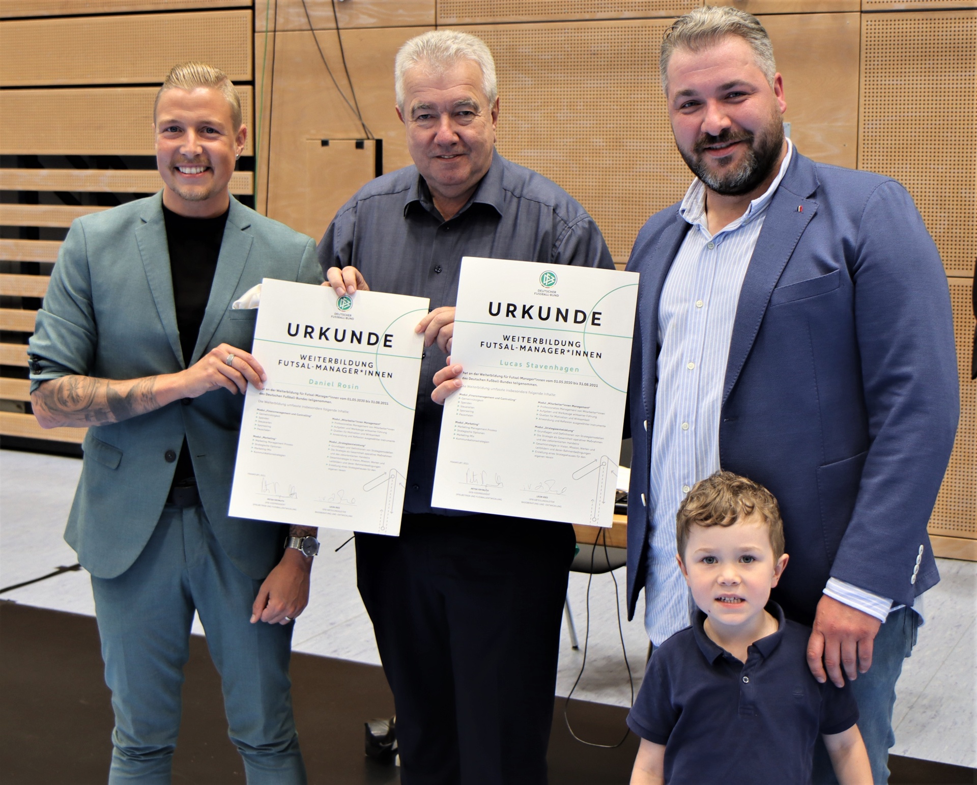 [{"clang_id":1,"value":"Vier Männer und ein Kind stehen lächelnd in einem Raum. Zwei Männer halten Urkunden in den Händen, vermutlich nach einer Weiterbildung im Futsal-Bereich."}]