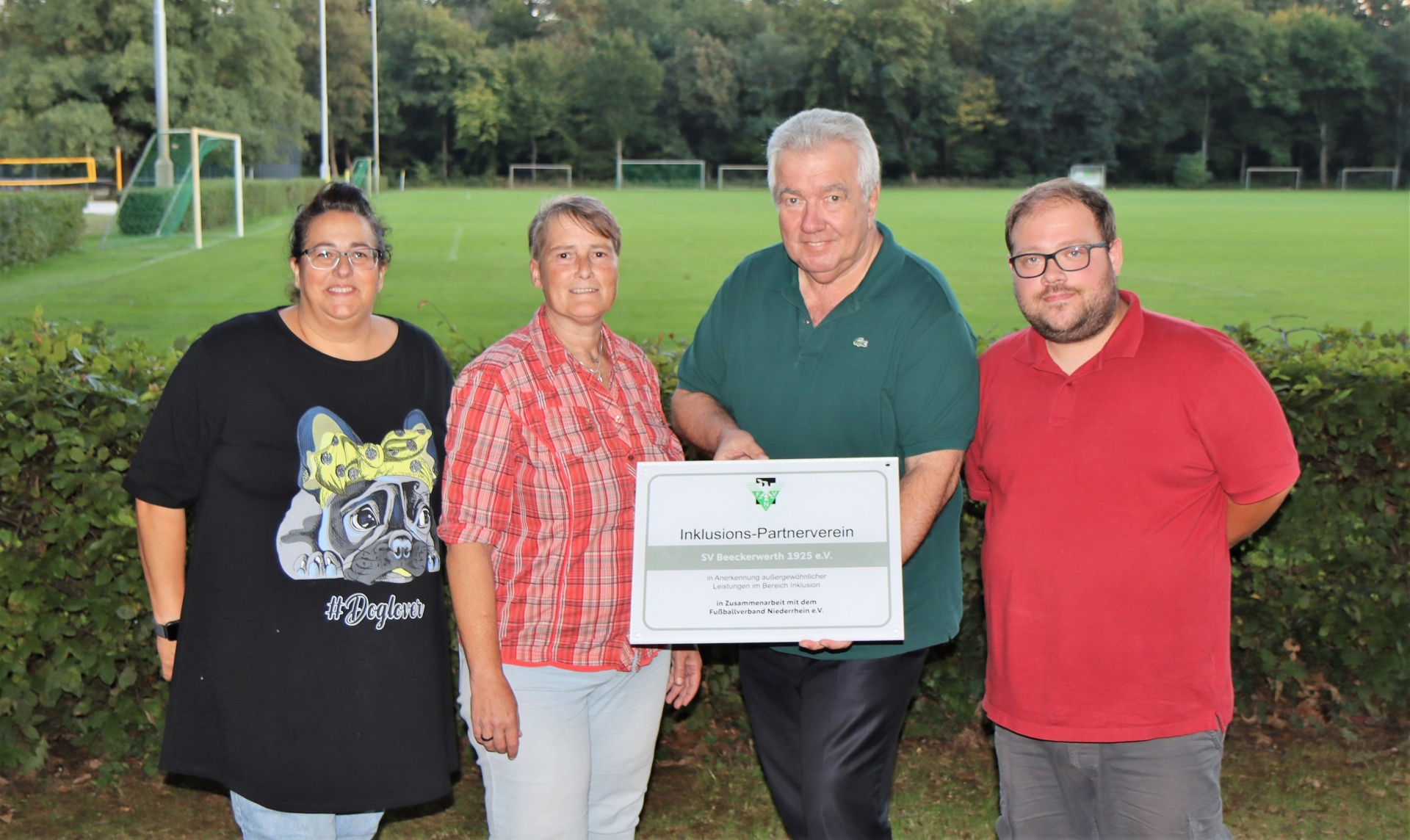 [{"clang_id":1,"value":"Vier Personen stehen vor einem Fußballplatz. Von links: Frau mit T-Shirt, Frau mit kariertem Hemd, Mann mit Polohemd und Mann mit braunem Hemd. Der Mann mit Polohemd hält eine Informationstafel."}]