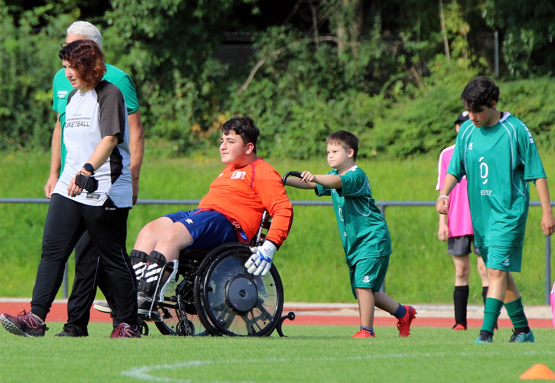 [{"clang_id":1,"value":"Sportplatz: Eine Person im Rollstuhl wird von einem Kind und einer erwachsenen Frau begleitet. Im Hintergrund weitere Kinder in Sportkleidung."}]
