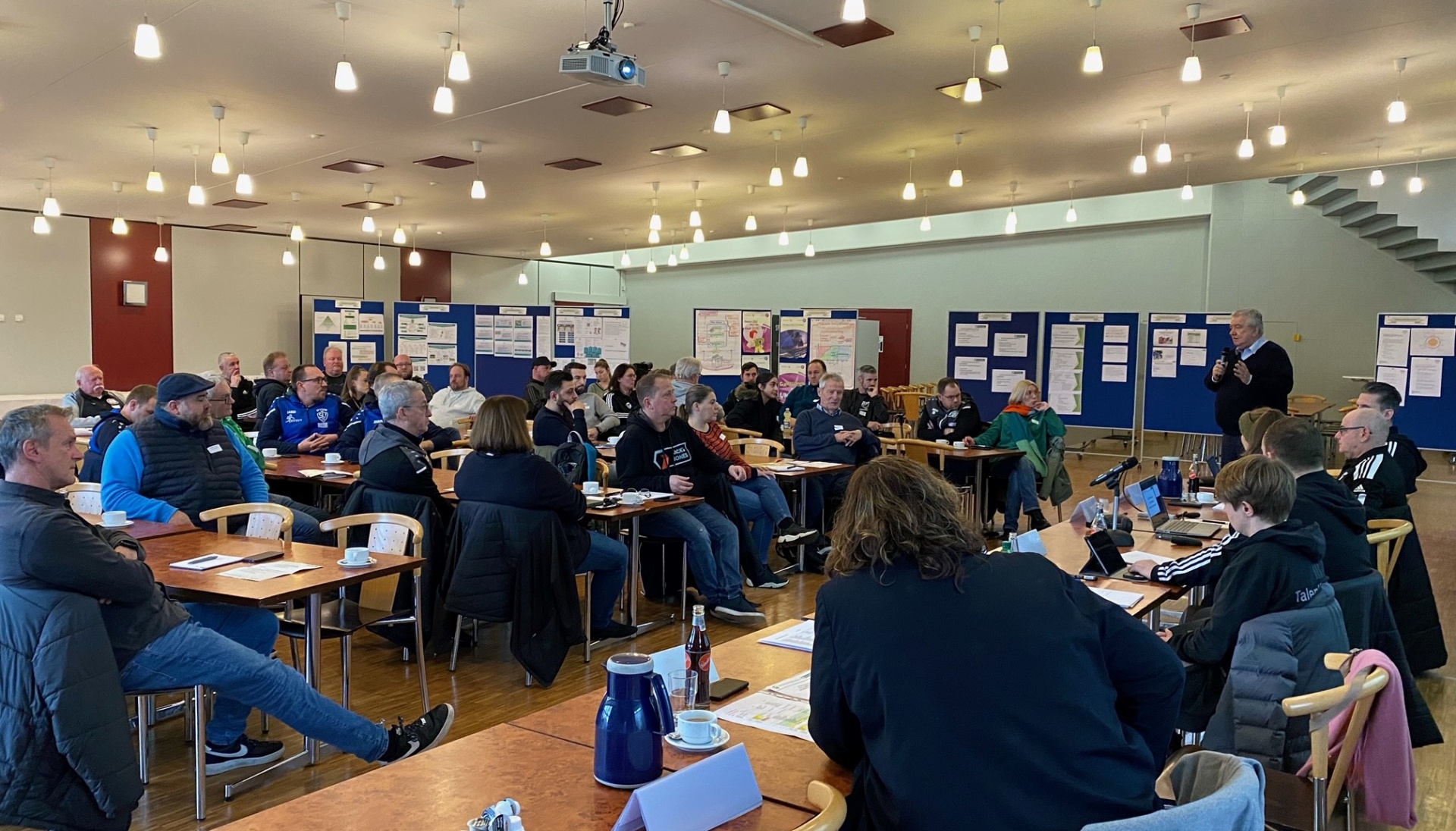 [{"clang_id":1,"value":"Großer Raum mit etwa 30 Personen bei einer Versammlung oder einem Meeting. Tische und Stühle sind verteilt aufgestellt, Wände mit Pinnwänden."}]