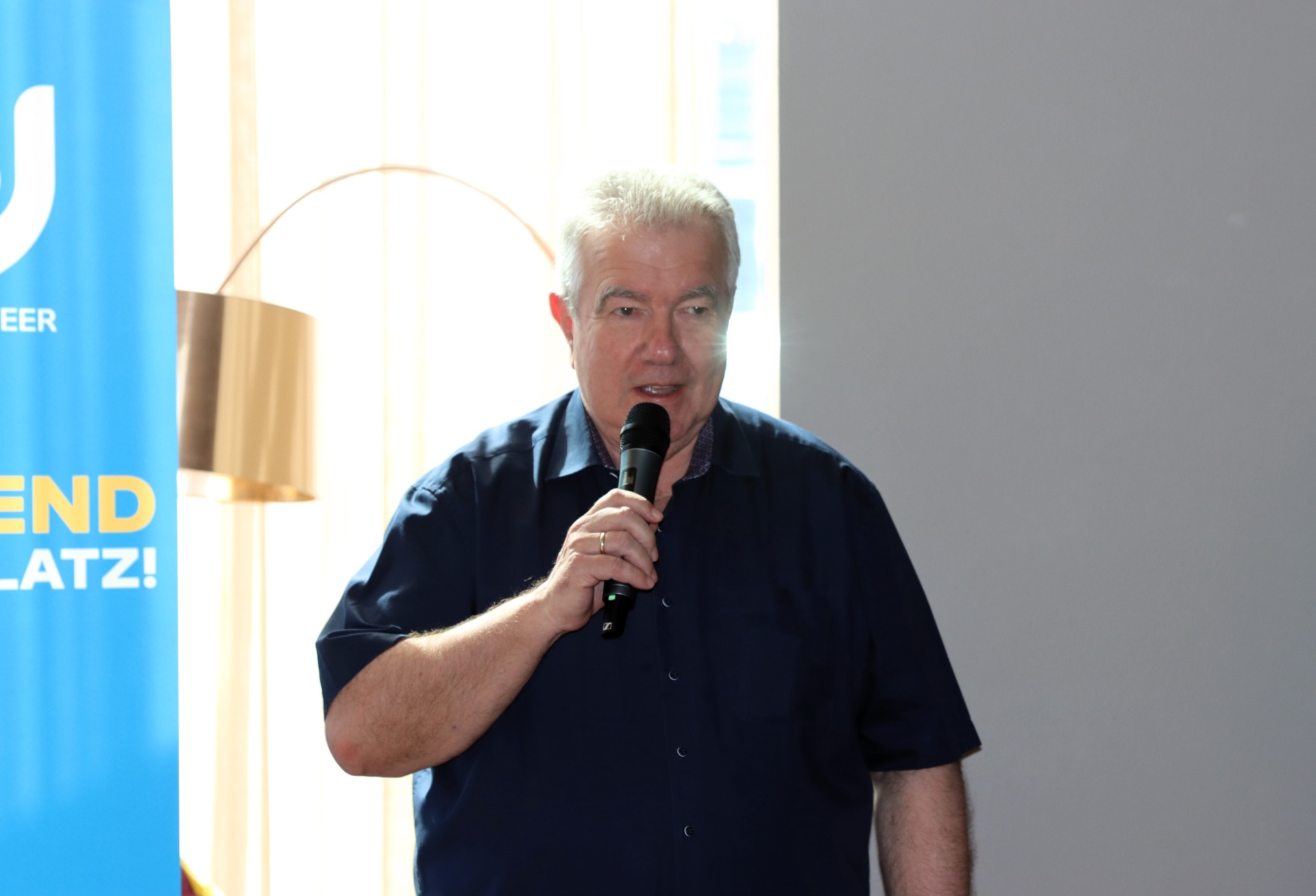 [{"clang_id":1,"value":"Mann mit Mikrofon spricht vor Publikum. Im Hintergrund ein Banner mit blauem Rand und dem Text \"ENDPLATZ!\". Großer Bogen im Hintergrund."}]