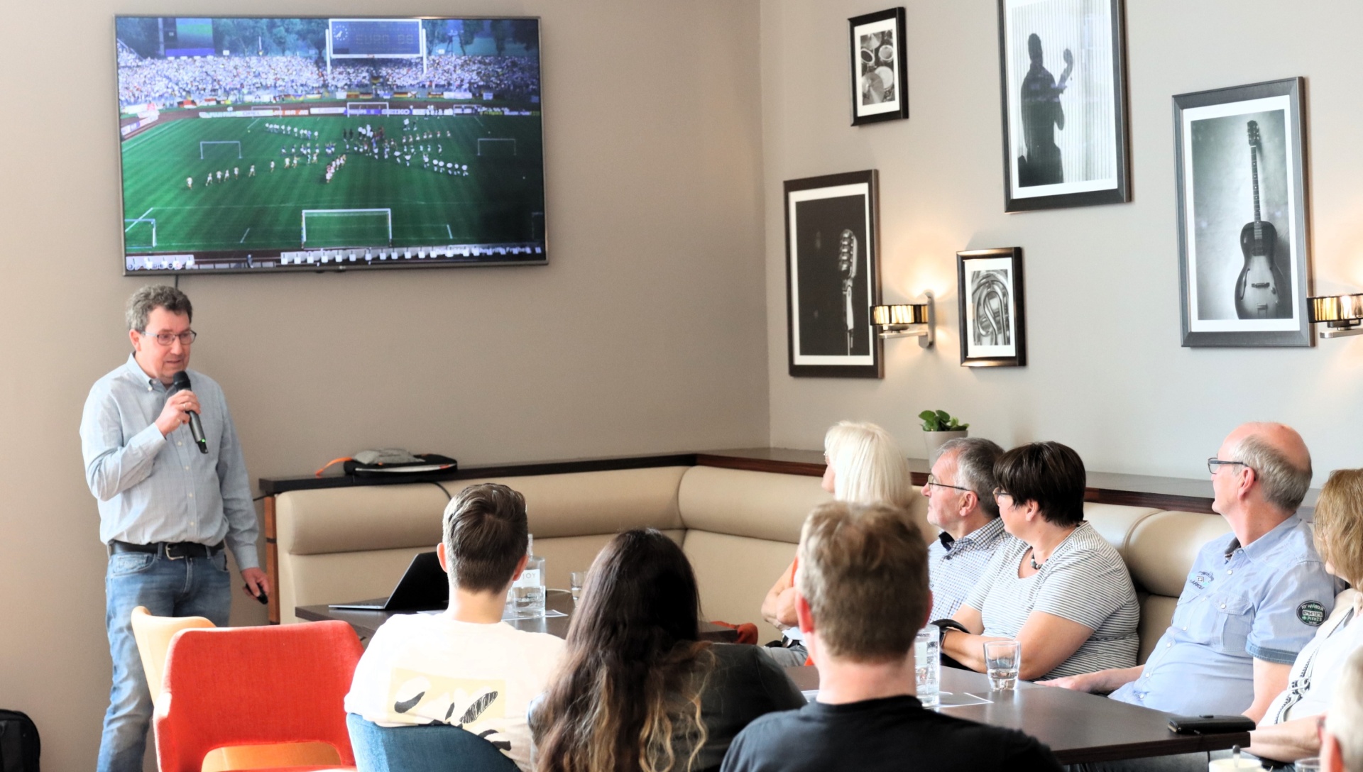 [{"clang_id":1,"value":"Mann mit Mikrofon spricht vor Gruppe sitzender Personen. Im Hintergrund eine große Leinwand mit Fußballspiel. Helle, moderne Raumgestaltung mit Sofas und Bildern an der Wand."}]