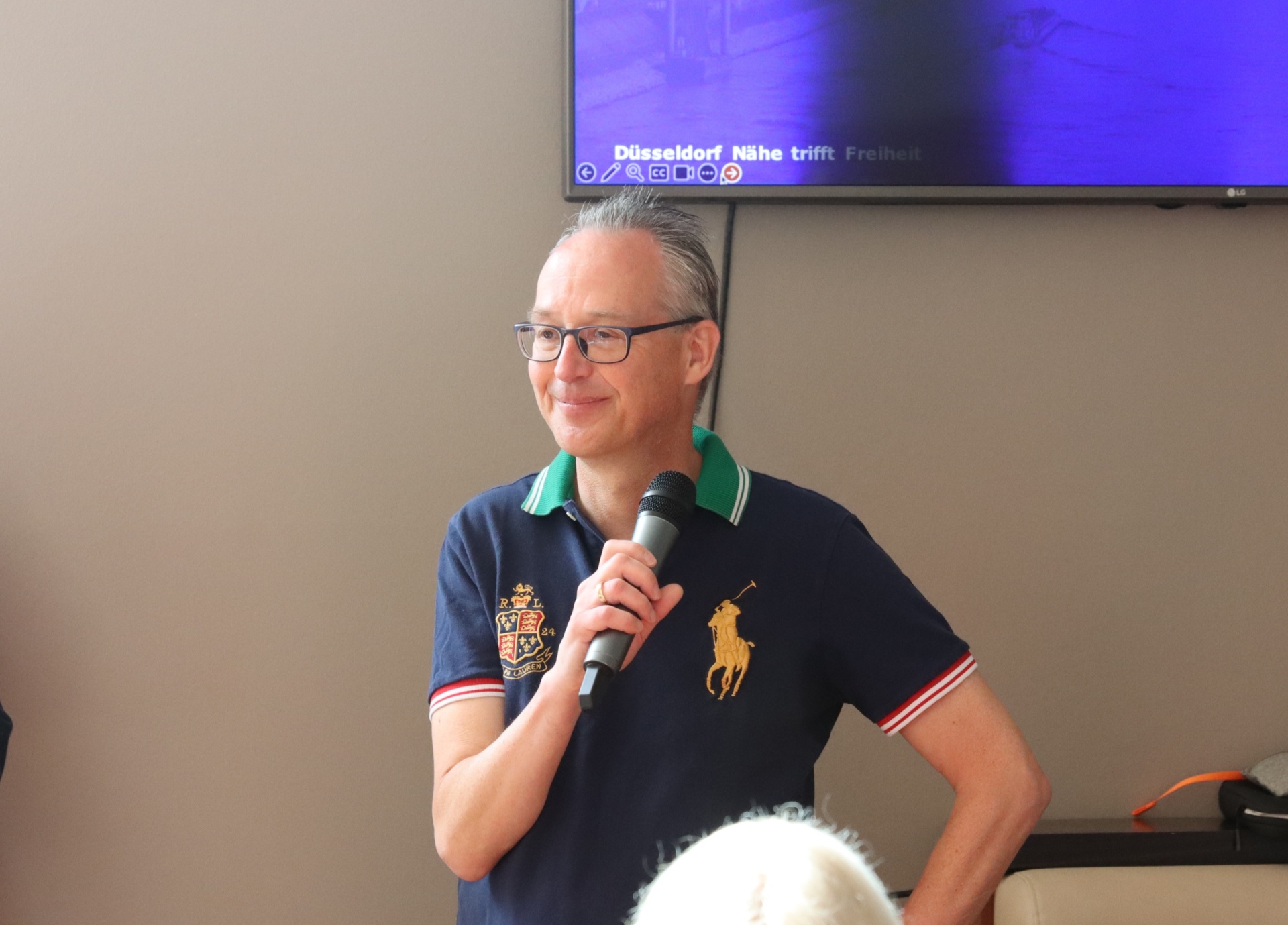 [{"clang_id":1,"value":"Mann mit Brille und Polo-Shirt hält Mikrofon. Im Hintergrund eine Leinwand mit Text \"Düsseldorf\"."}]