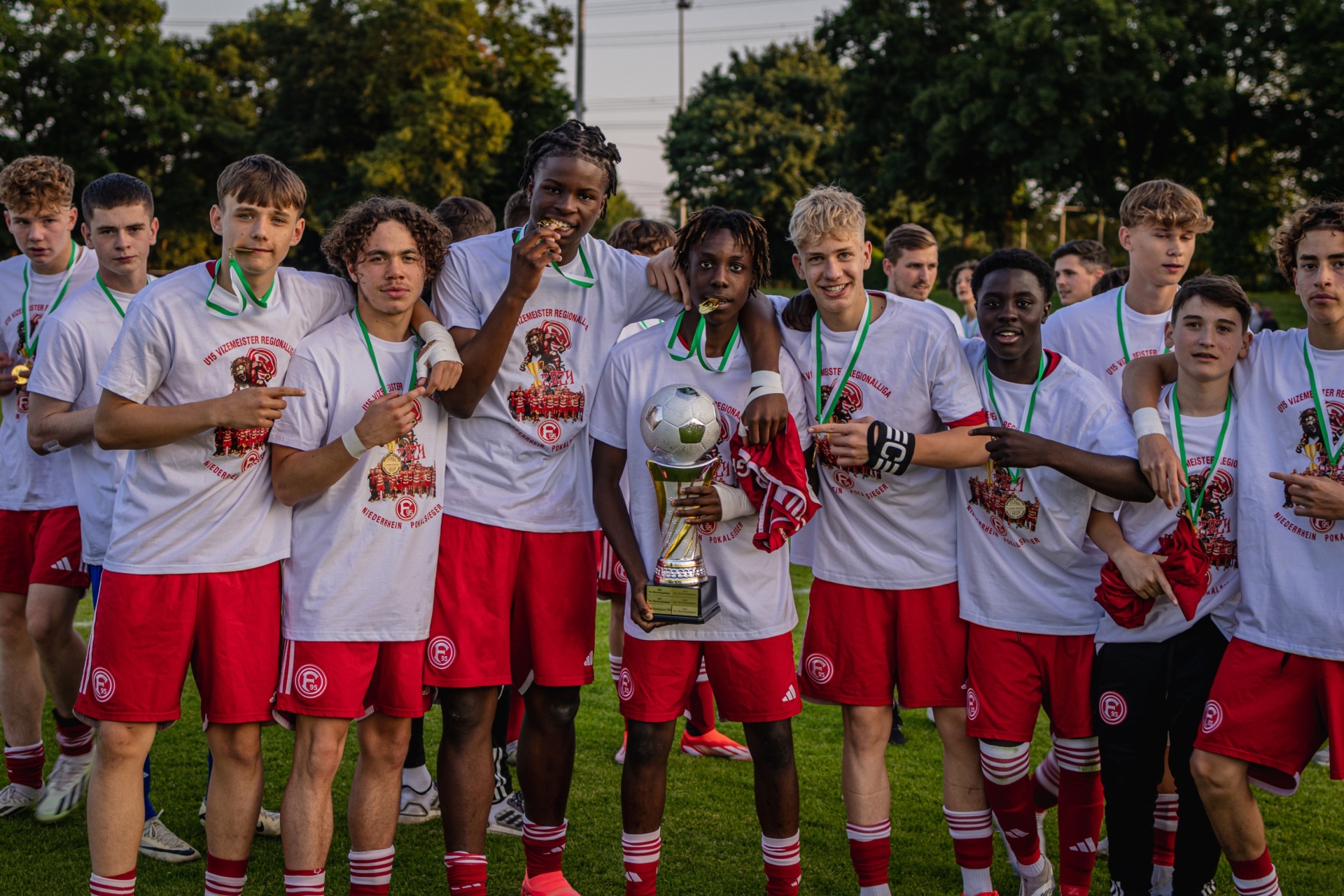 [{"clang_id":1,"value":"Fußballmannschaft in rot-weißen Trikots feiert Sieg mit Pokal und Medaillen im Freien."}]