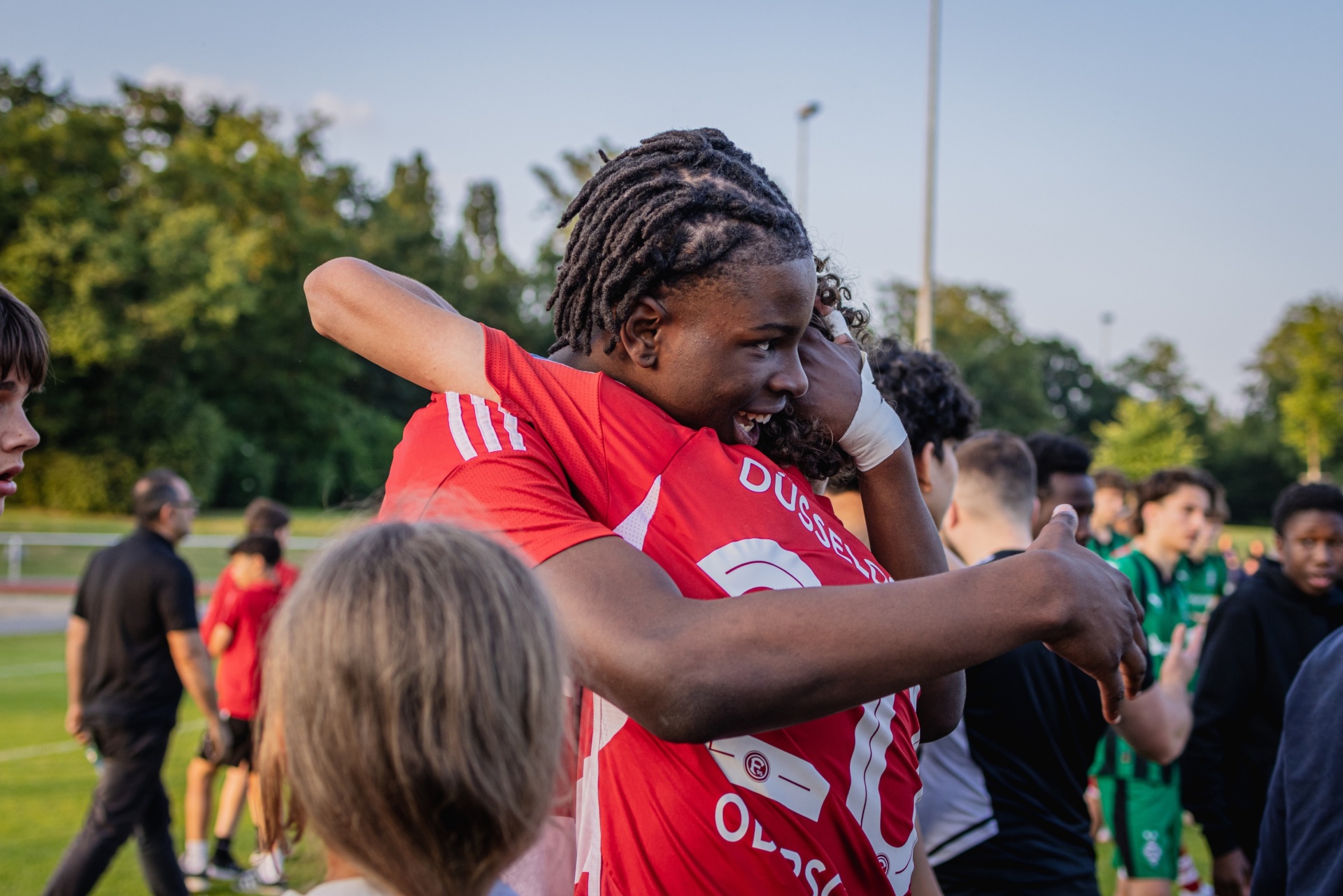 [{"clang_id":1,"value":"Zwei Fußballspieler in roten Trikots umarmen sich jubelnd auf einem grünen Rasenfeld. Im Hintergrund weitere Spieler und Zuschauer."}]