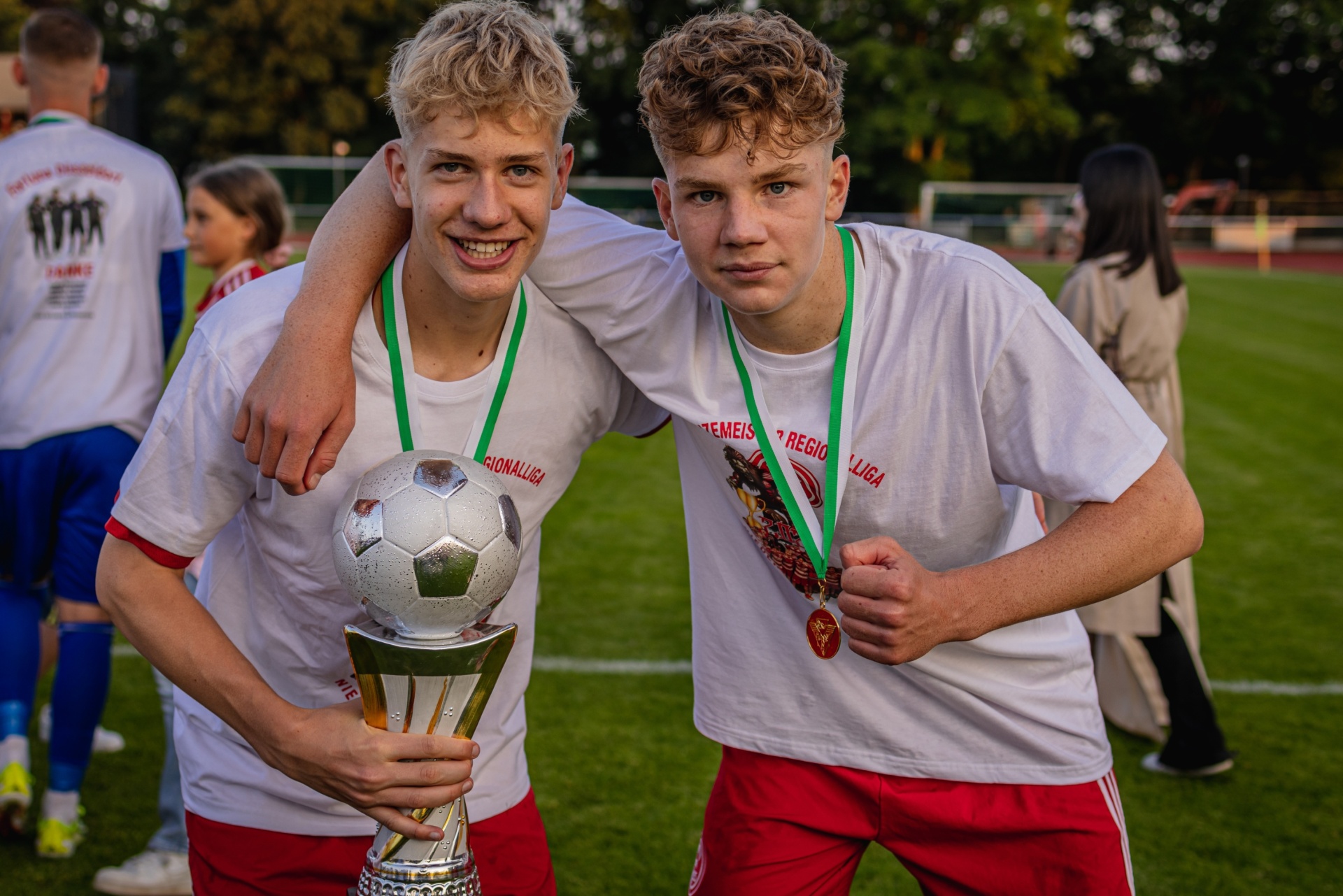 [{"clang_id":1,"value":"Zwei junge Fussballspieler mit Medaillen und Pokal umarmen sich auf einem Fussballfeld. Im Hintergrund weitere Spieler und Zuschauer."}]