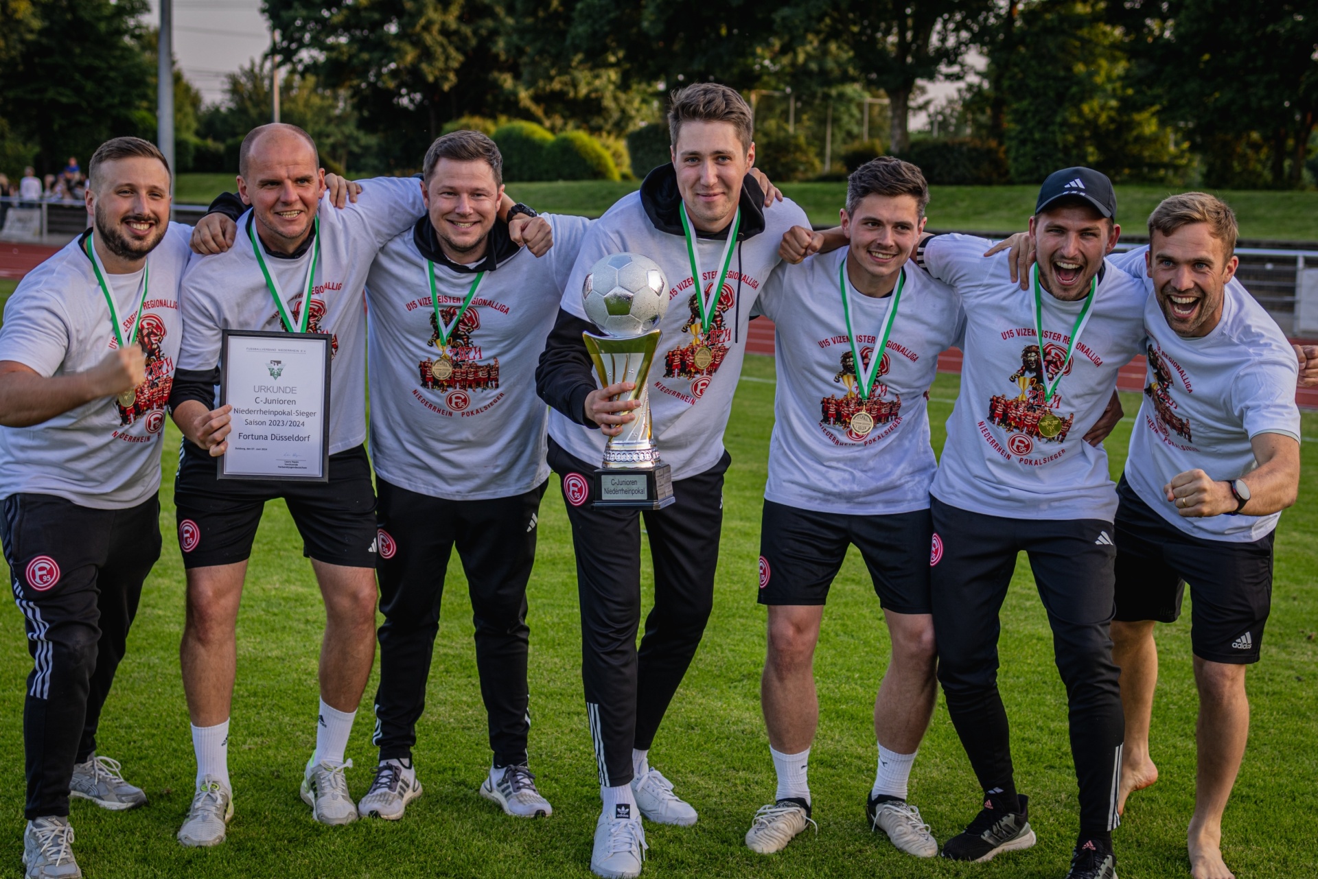 [{"clang_id":1,"value":"Fußballmannschaft mit Pokal und Urkunde. Neun Männer stehen auf einem Fußballfeld, lächeln und halten den Pokal und die Urkunde hoch. Sie tragen rote T-Shirts und Sportkleidung."}]