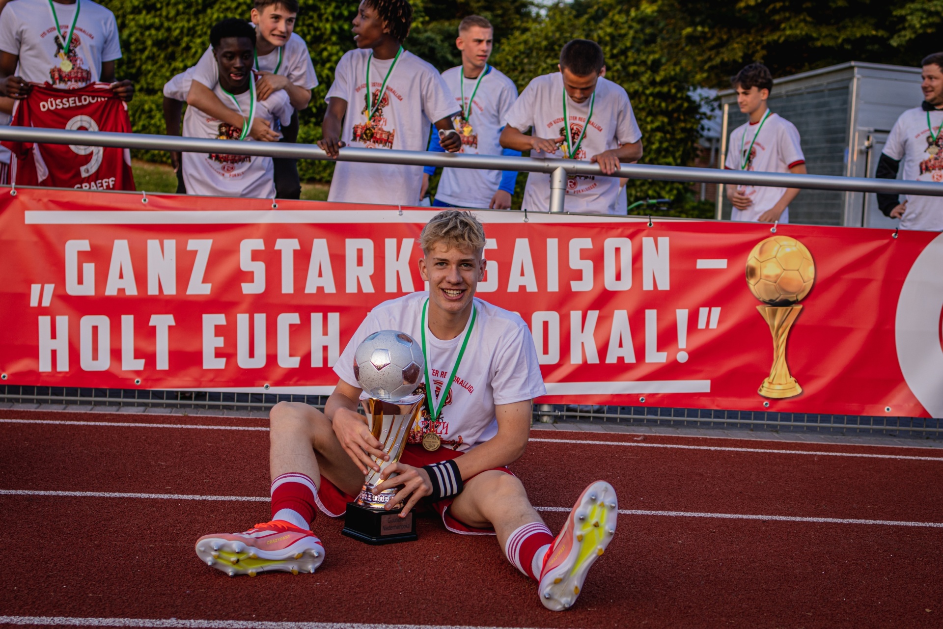 [{"clang_id":1,"value":"Junge/r Fußballspieler/in sitzt lächelnd mit zwei Pokalen auf einer Laufbahn. Im Hintergrund steht die Mannschaft mit Medaillen, ein Banner mit Schrift und Fußball-Motiv."}]