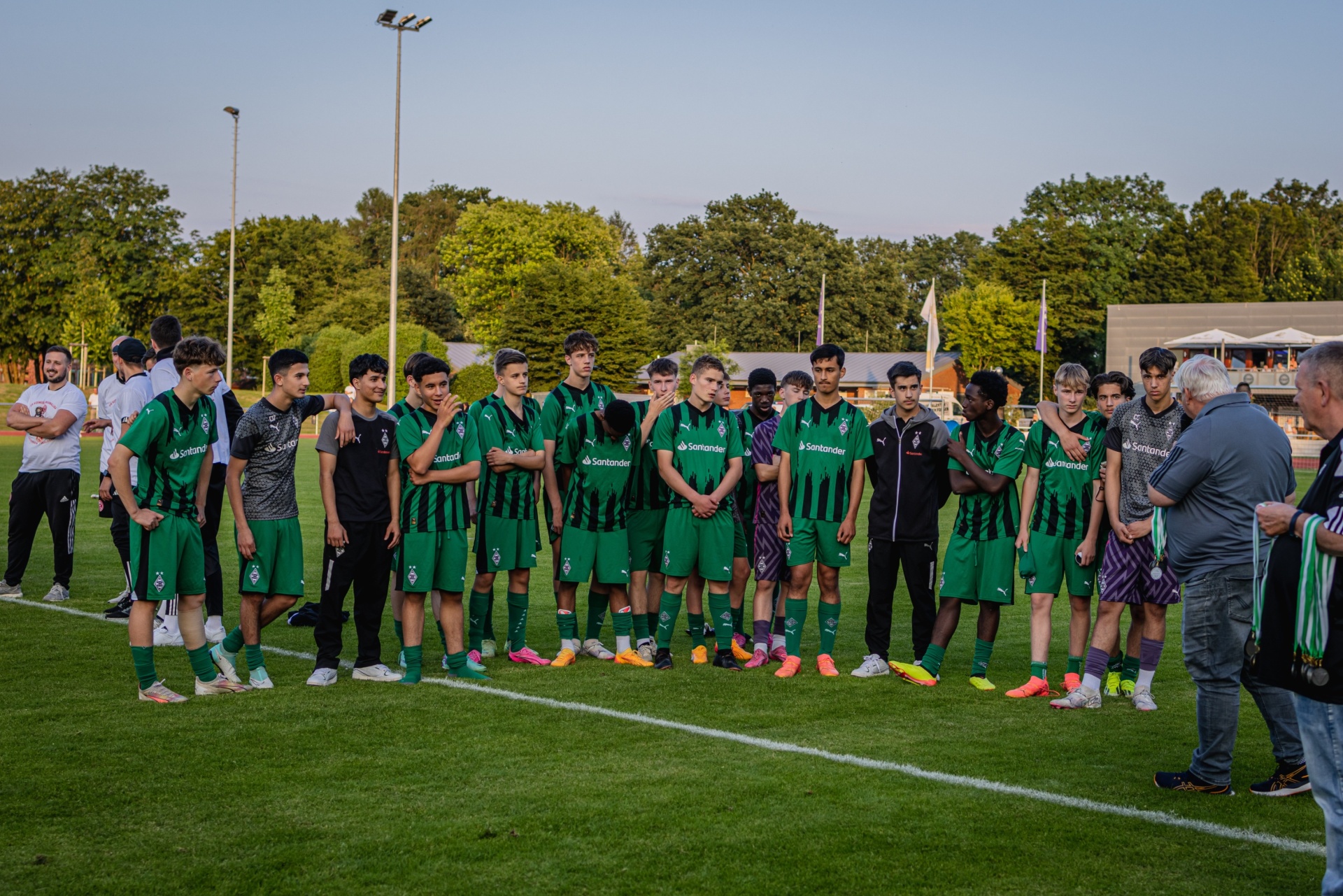 [{"clang_id":1,"value":"Fußballmannschaft in grünen Trikots und Shorts steht auf einem Fußballfeld. Trainer und Zuschauer im Hintergrund. Bäume säumen das Spielfeld."}]