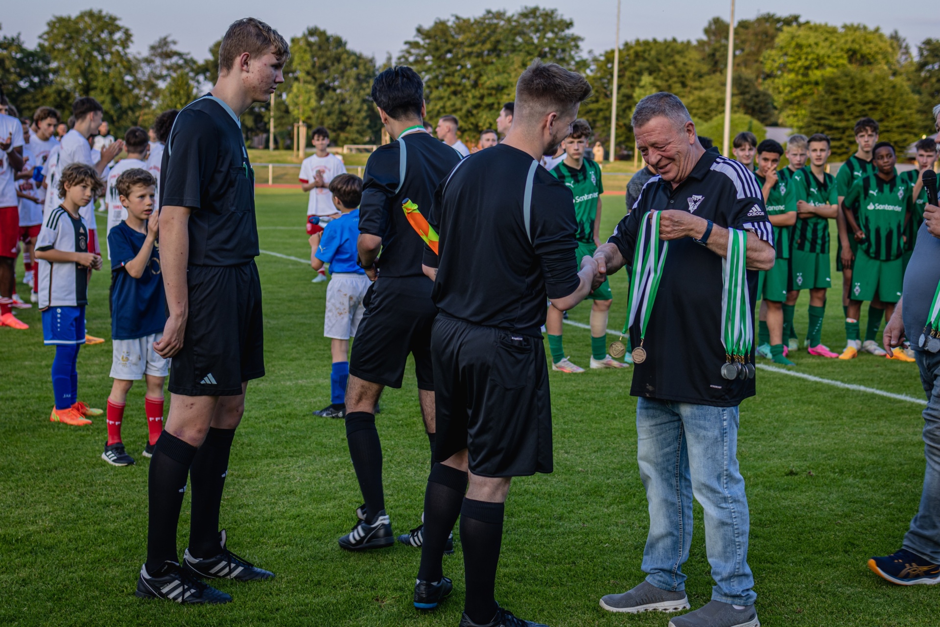 [{"clang_id":1,"value":"Fußballspieler und Trainer erhalten Medaillen auf einem grünen Spielfeld. Im Hintergrund stehen Zuschauer und Bäume."}]
