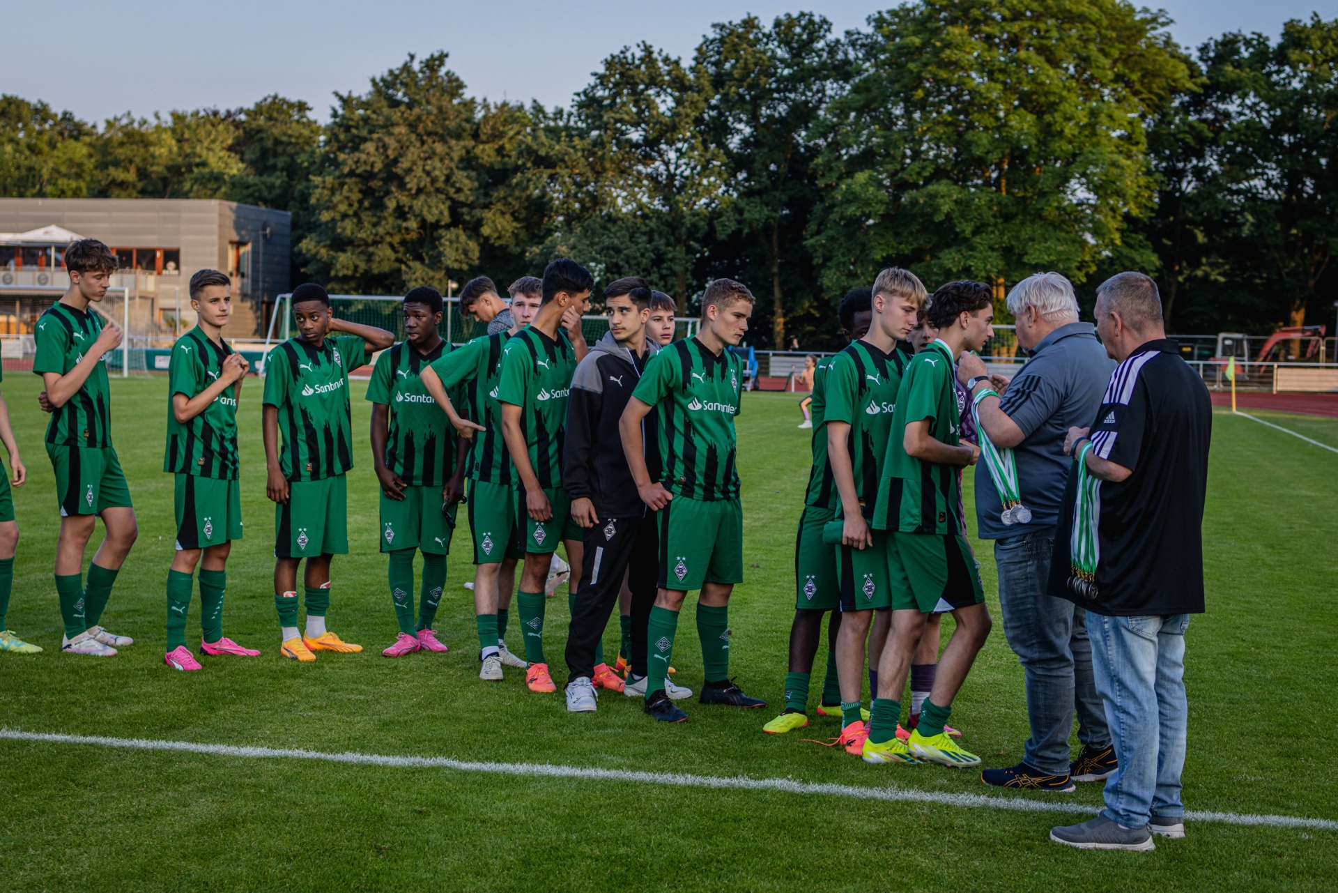 [{"clang_id":1,"value":"Fußballmannschaft in grünen Trikots und der Trainer stehen auf einem Rasenplatz. Alle blicken auf den Trainer, der ein Tablet hält. Bäume im Hintergrund."}]