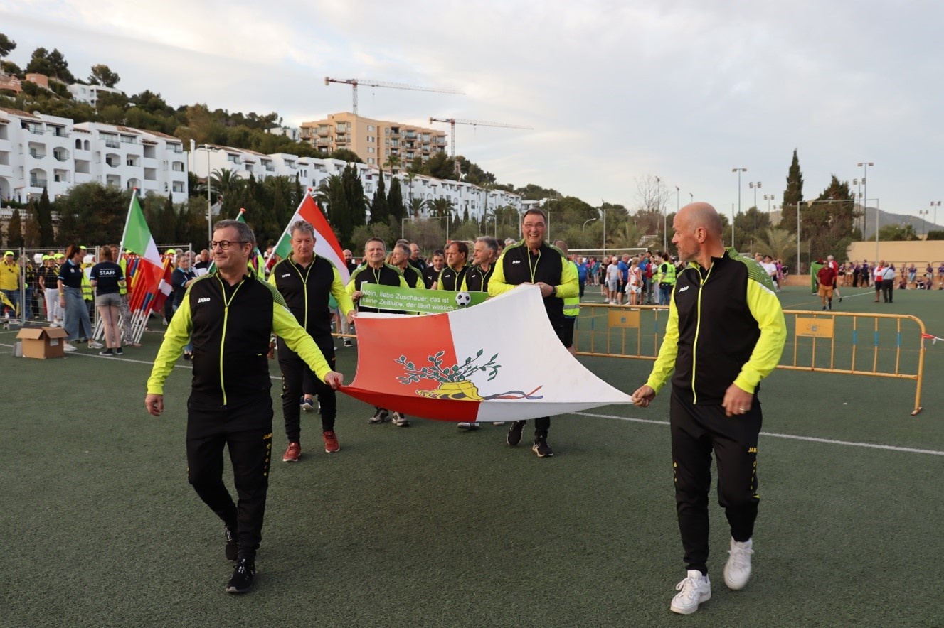 [{"clang_id":1,"value":"Fußballfeld mit einer Gruppe Personen, die Flaggen tragen. Im Hintergrund Gebäude und Bäume bei Tageslicht."}]