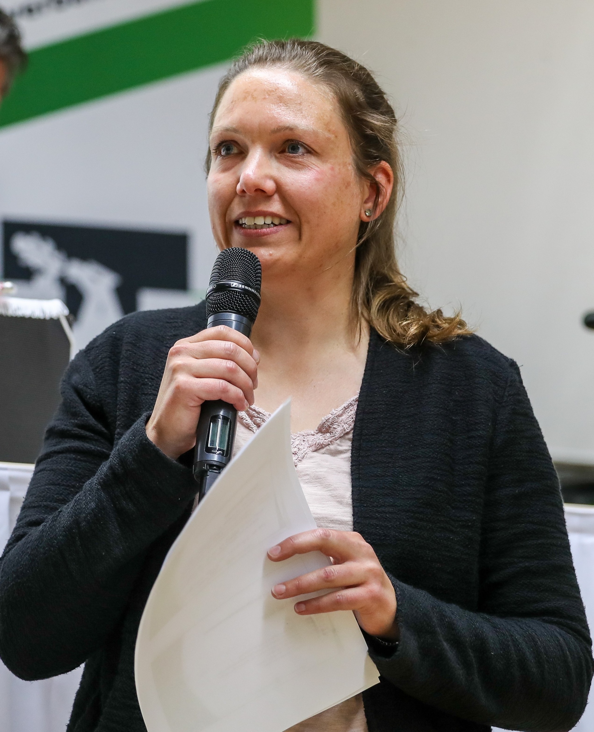 [{"clang_id":1,"value":"Frau mit braunen Haaren hält Mikrofon und Papier. Sie lächelt und trägt eine graue Strickjacke über einem beigen Top. Im Hintergrund verschwommene Personen und eine grüne Wand."}]