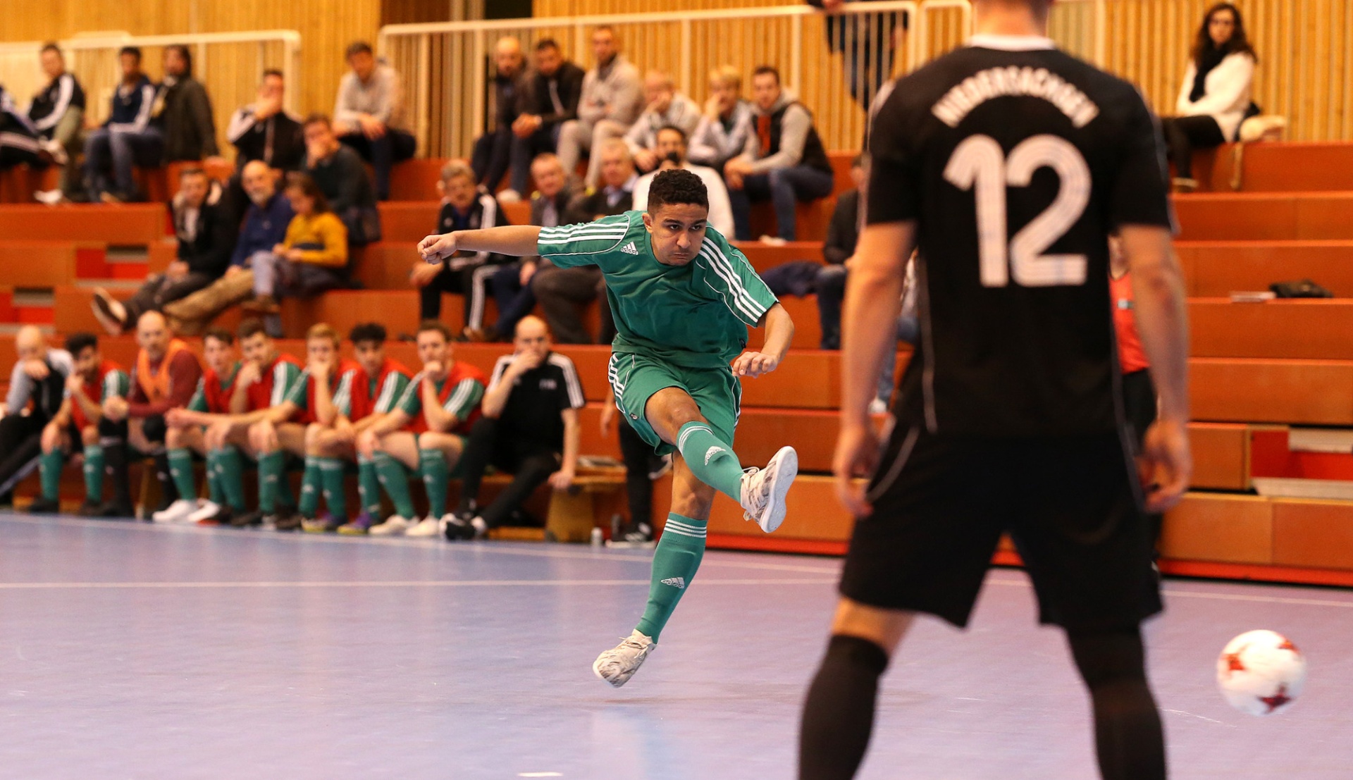 [{"clang_id":1,"value":"Futsalspieler in grünen Trikots schießt auf das Tor. Im Hintergrund Zuschauerreihen und ein Spieler in Schwarz mit der Nummer 12."}]