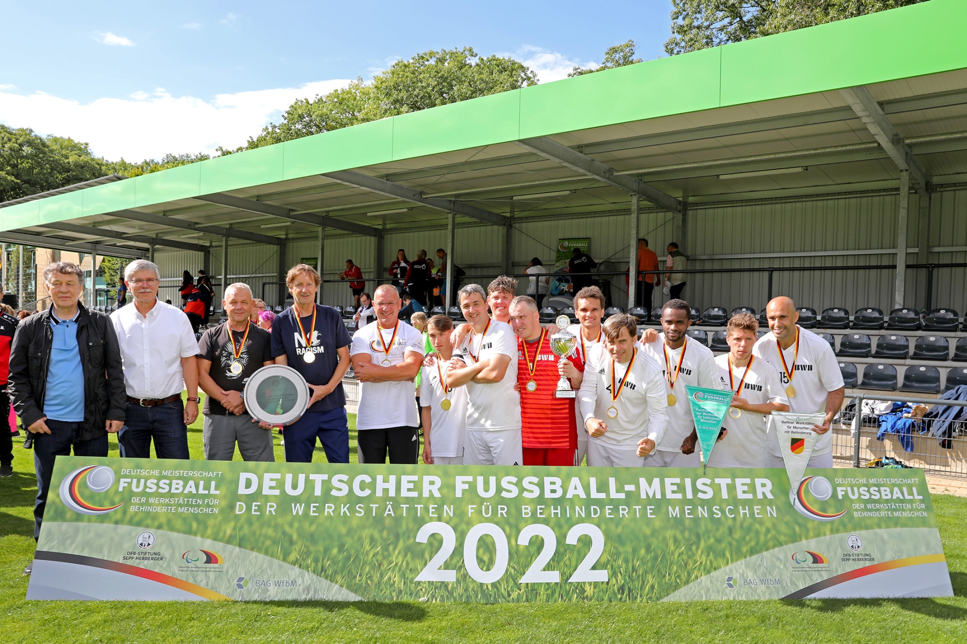 [{"clang_id":1,"value":"Fußballmannschaft mit Medaillen und Pokal vor Banner mit Aufschrift \"Deutscher Fußball-Meister der Werkstätten für behinderte Menschen 2022\". Stadionhintergrund."}]