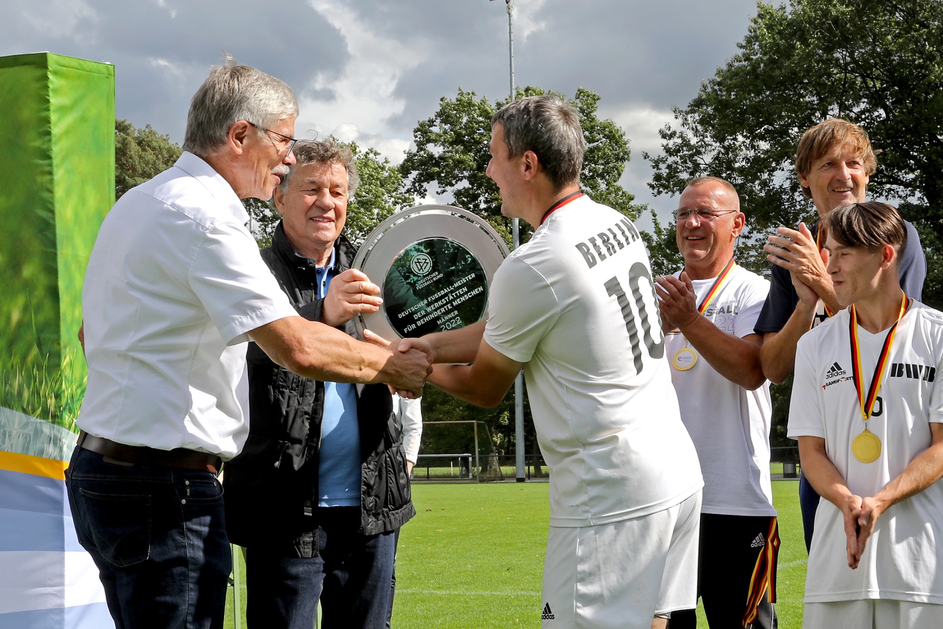 [{"clang_id":1,"value":"Gruppe posiert bei Preisverleihung. Mann in Fußballtrikot mit Nummer 10 erhält Auszeichnung von zwei Herren. Hintergrund: Grünfläche und Zuschauer."}]