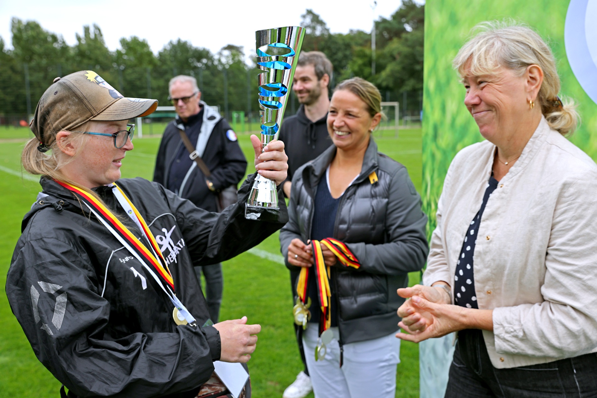 [{"clang_id":1,"value":"Eine Frau mit Kappe hält einen Pokal hoch, umgeben von drei weiteren Personen, die klatschen oder zusehen. Im Hintergrund ein Fußballfeld und Bäume."}]
