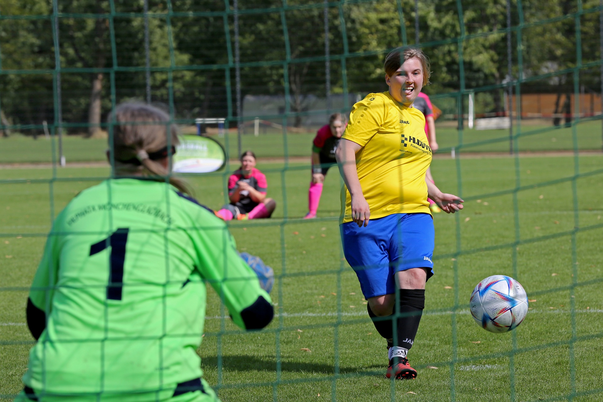 [{"clang_id":1,"value":"Fußballspiel: Spielerin in gelbem Trikot schießt auf das Tor. Im Tor steht die Torhüterin in grünem Outfit. Hintergrund: Zuschauer und Bäume."}]