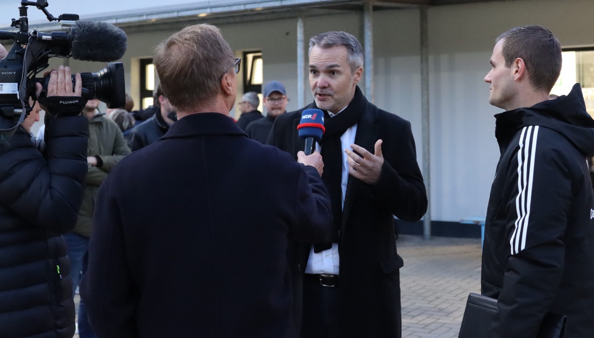 [{"clang_id":1,"value":"Interview mit einem Mann in schwarzem Mantel vor laufender Kamera. Im Hintergrund weitere Personen und ein Gebäude."}]