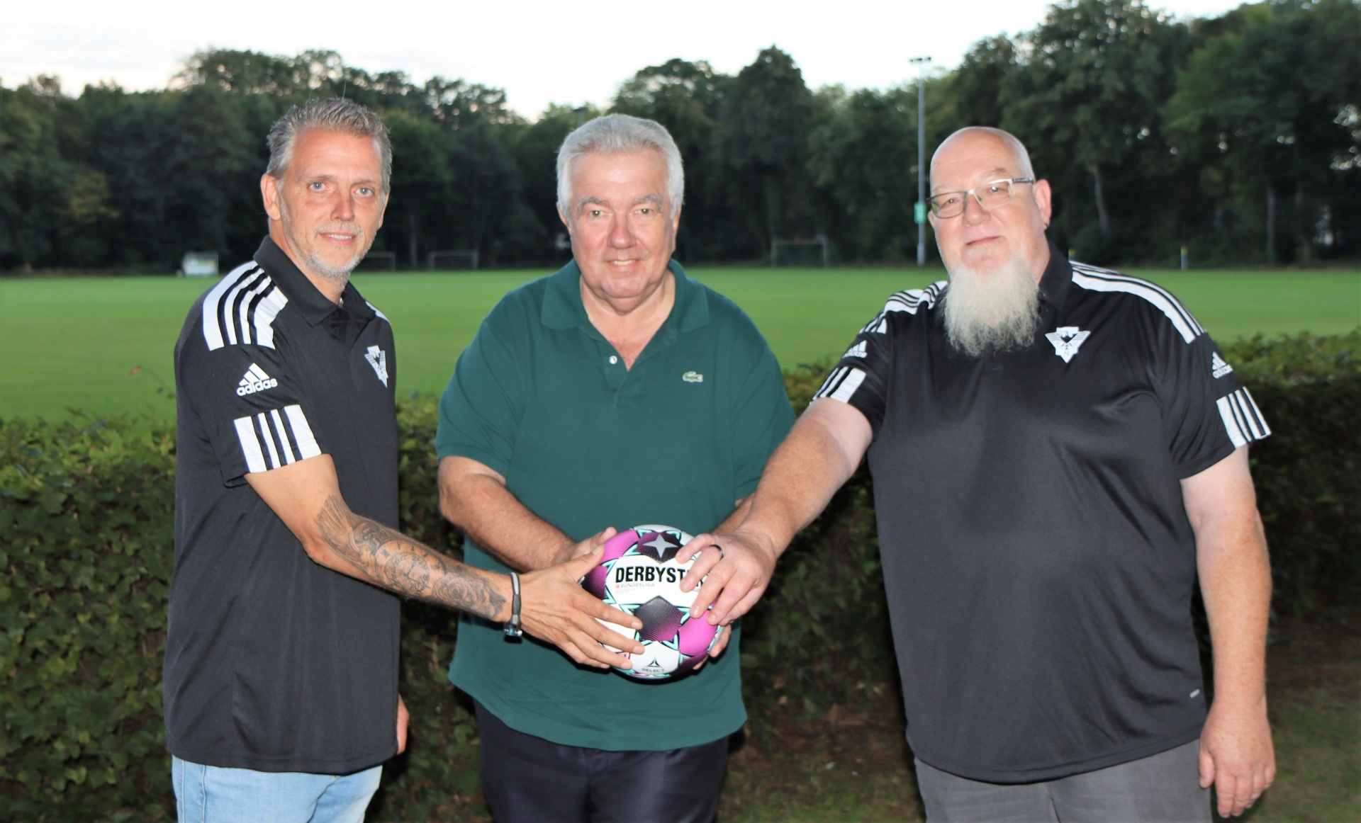 [{"clang_id":1,"value":"Drei Männer stehen auf einer Grünfläche vor einem Fußballfeld. Der Mittlere hält einen pinkfarbenen Fußball mit der Aufschrift \"DERBYST\"."}]