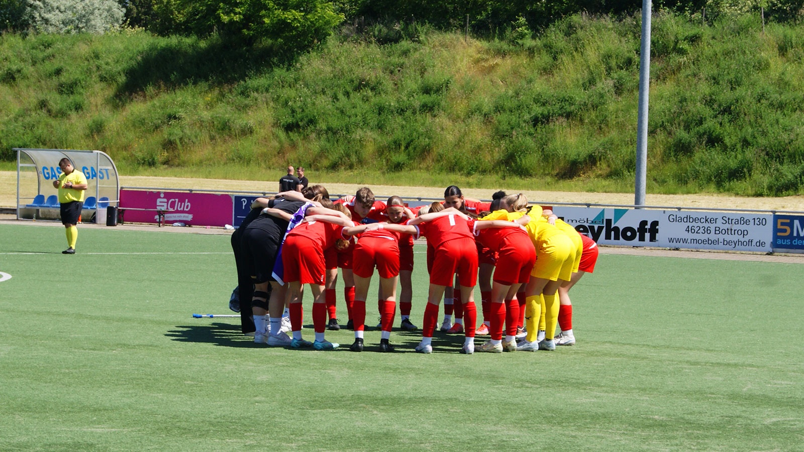 [{"clang_id":1,"value":"Fußballspielerinnen stehen im Kreis auf grünem Rasen, Arme ineinander gelegt. Im Hintergrund eine Zuschauertribüne und Werbebanner."}]
