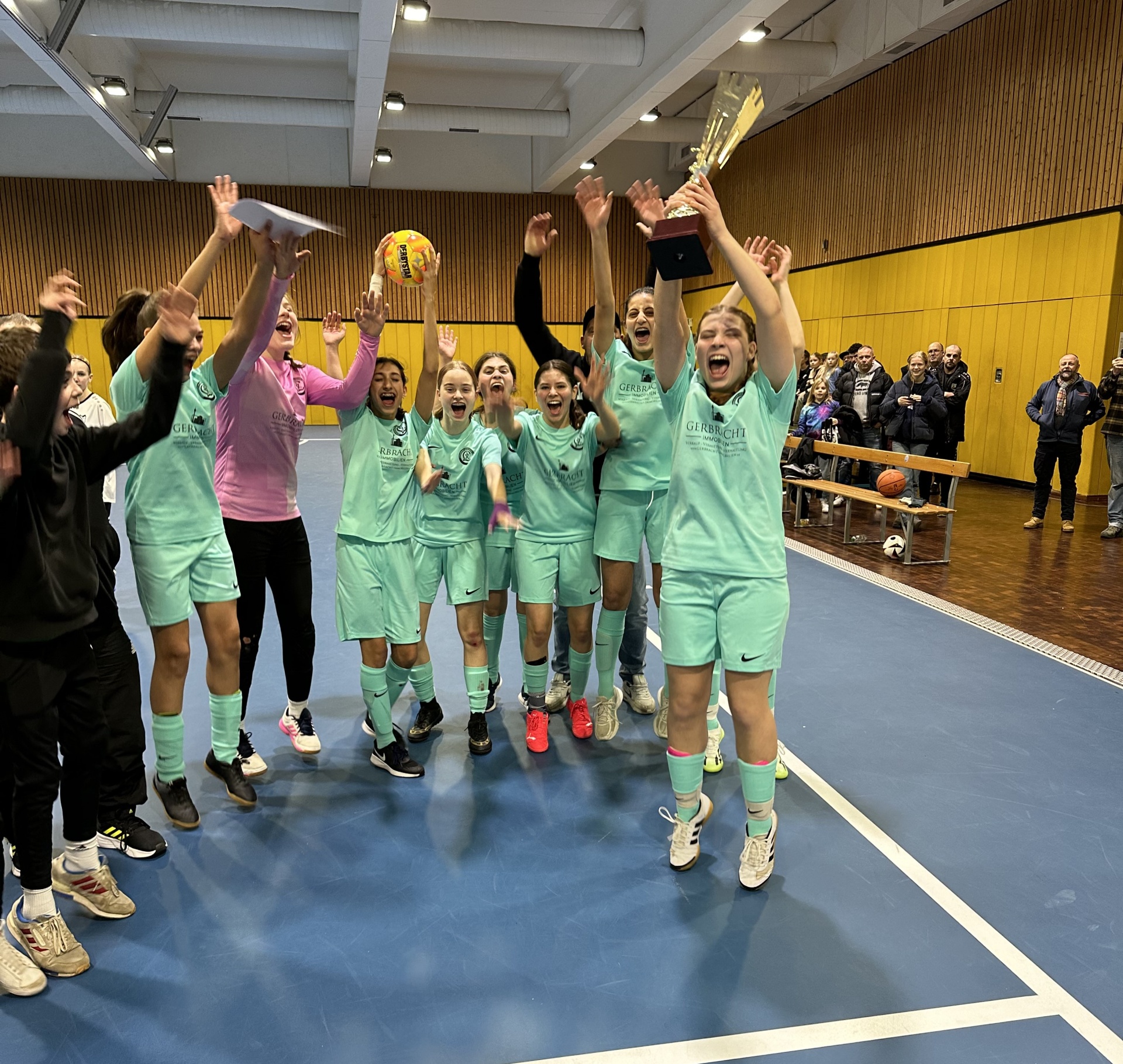 [{"clang_id":1,"value":"Basketballteam in hellgrünen Trikots feiert Sieg mit Pokalen und Konfetti in einer Sporthalle. Zuschauer im Hintergrund."}]
