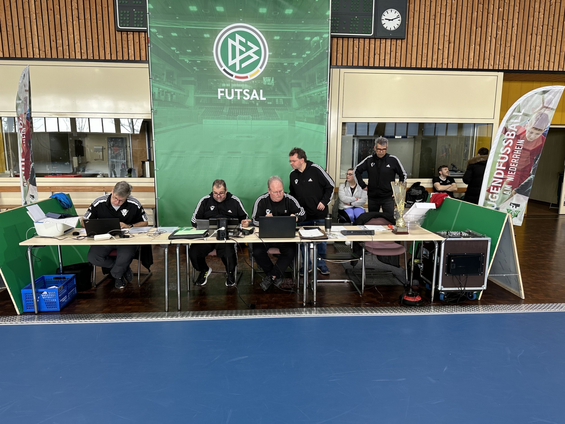 [{"clang_id":1,"value":"Futsal-Halle mit Tischreihe für Offizielle. Vier Personen sitzen an Laptops, weitere stehen und beobachten. Im Hintergrund eine große Futsal-Stadiongrafik."}]