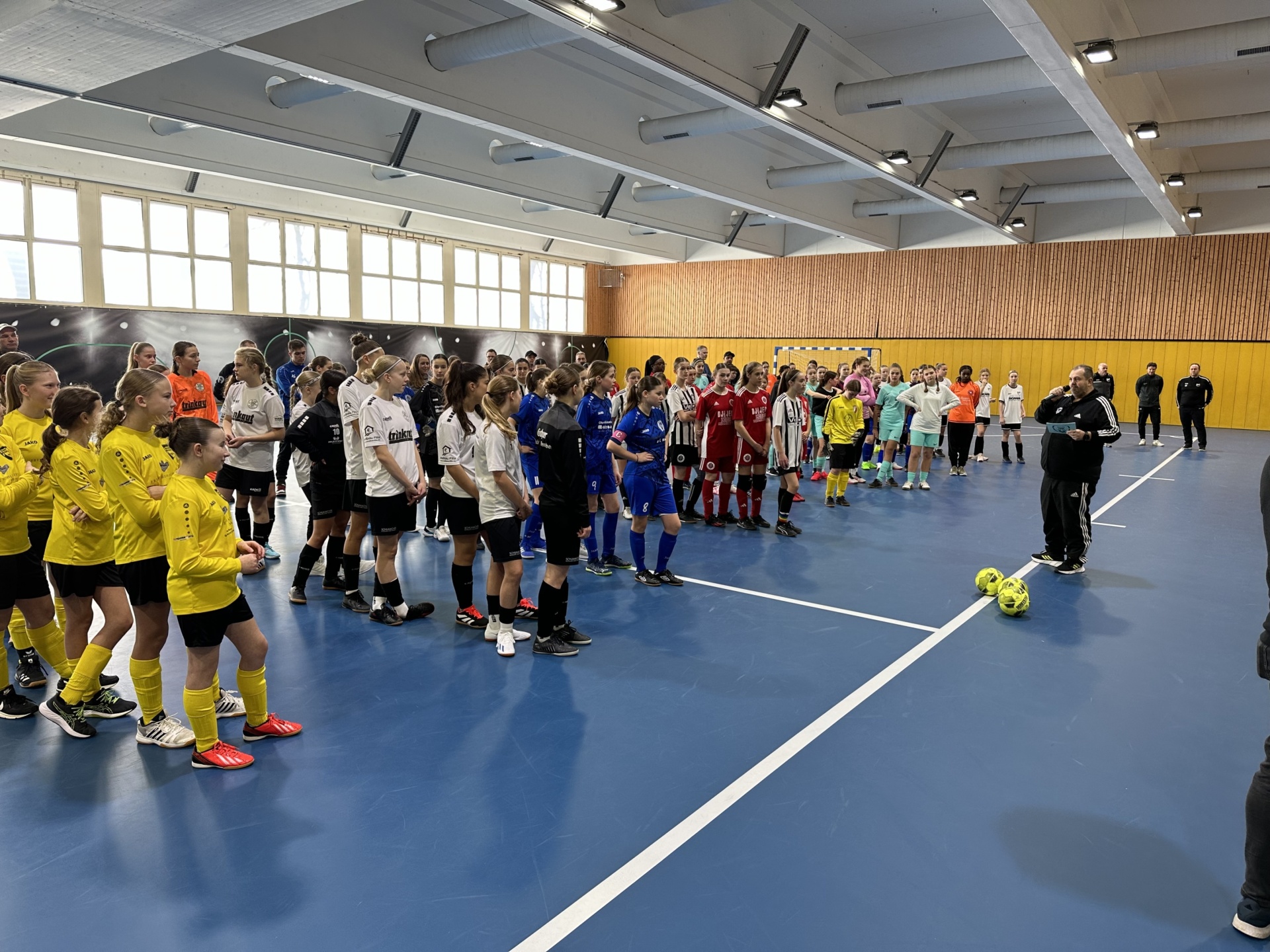 [{"clang_id":1,"value":"Große Gruppe Jugendlicher in Sportkleidung steht in einer Sporthalle. Ein Trainer gestikuliert, einige Bälle liegen am Boden."}]