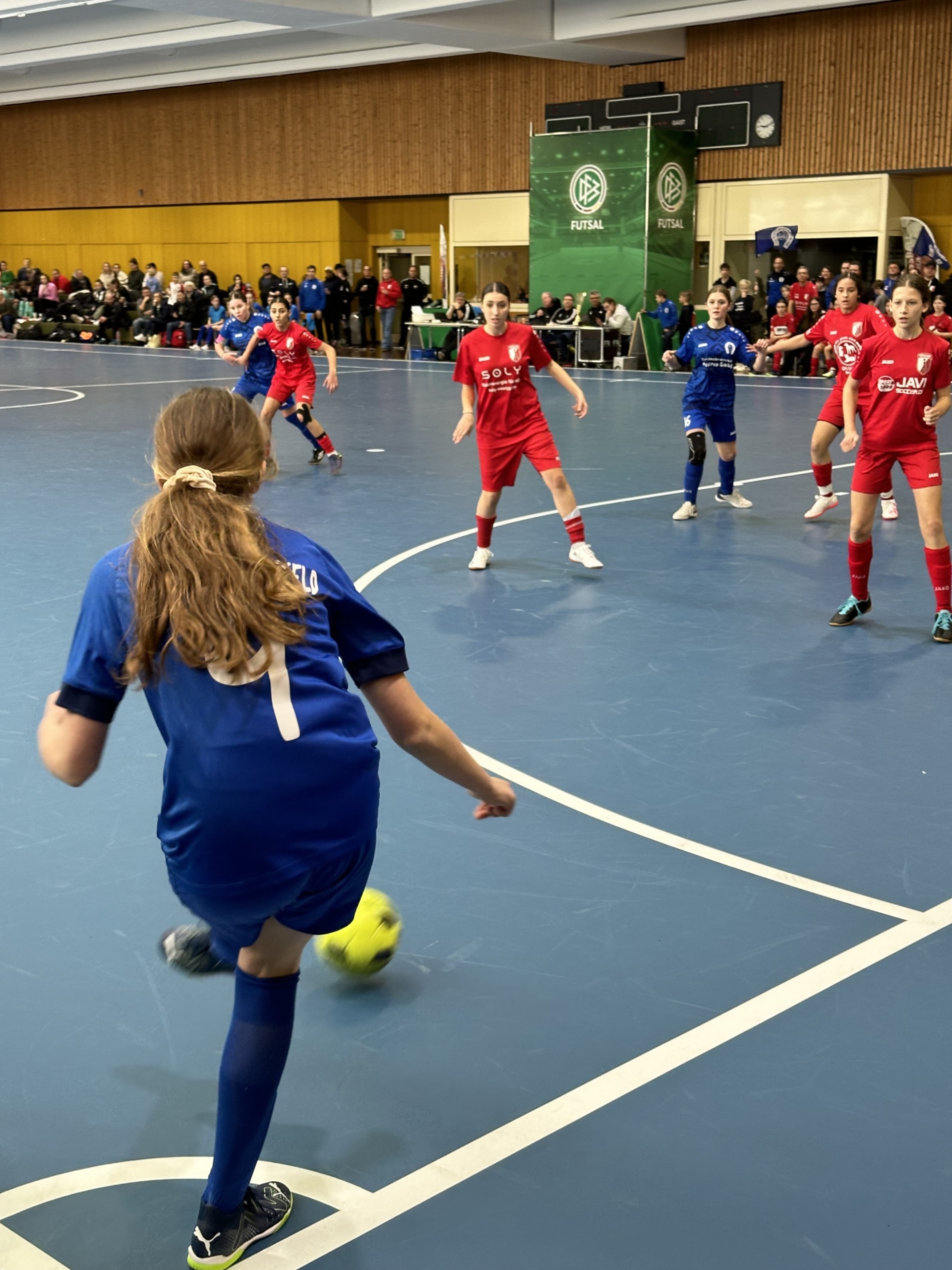 [{"clang_id":1,"value":"Futsalspiel in Sporthalle. Eine Spielerin mit blauem Trikot schießt auf den Ball, umgeben von weiteren Spielerinnen in blau und rot. Zuschauer auf der Empore."}]