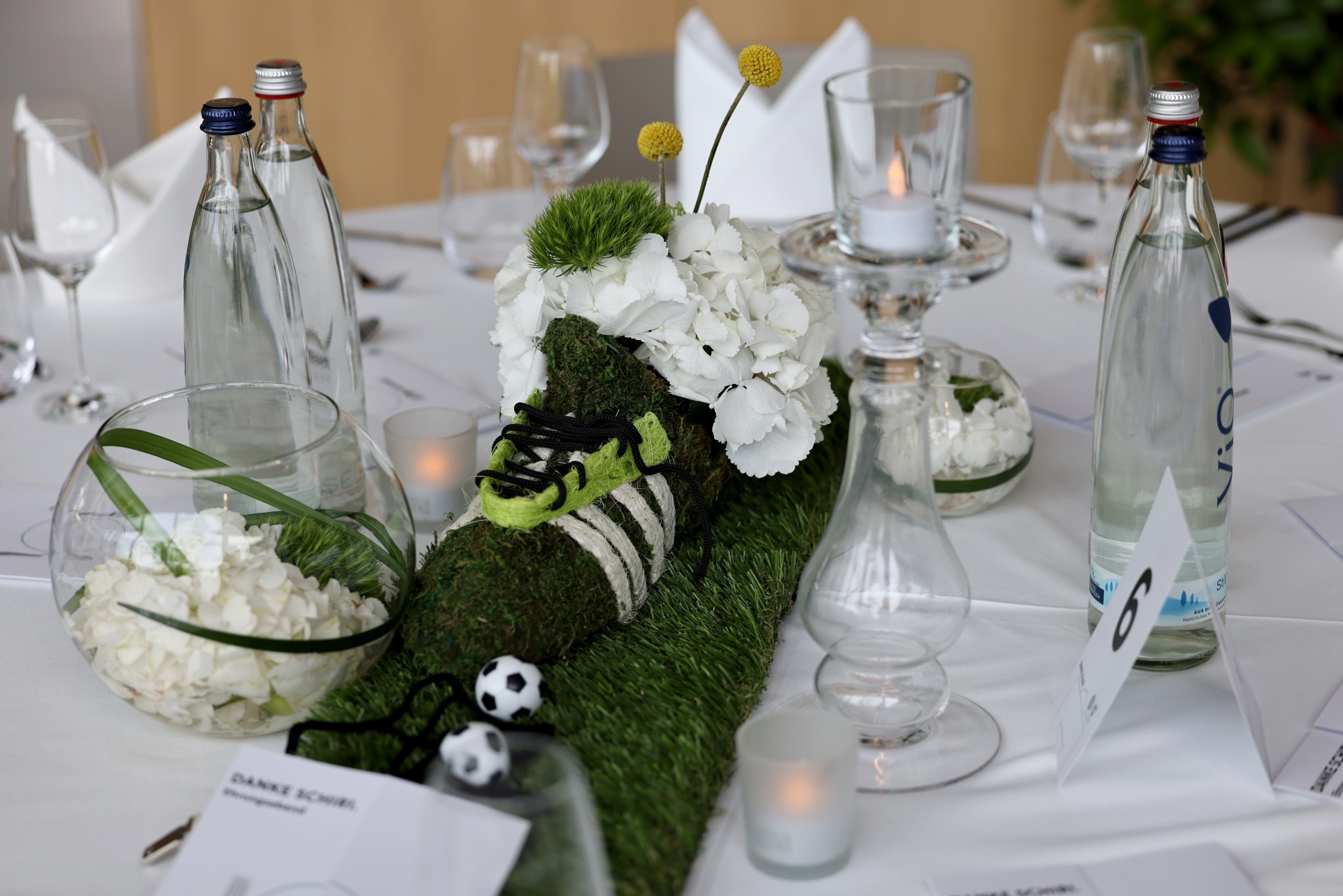 [{"clang_id":1,"value":"Tischdekoration mit Fußball-Motiv: Grasgrüne Tischdeko, Fußballschuh, Fußbälle, weiße Blumen, Kerzen, Wasserflaschen und Tischkarten auf weißer Tischdecke."}]