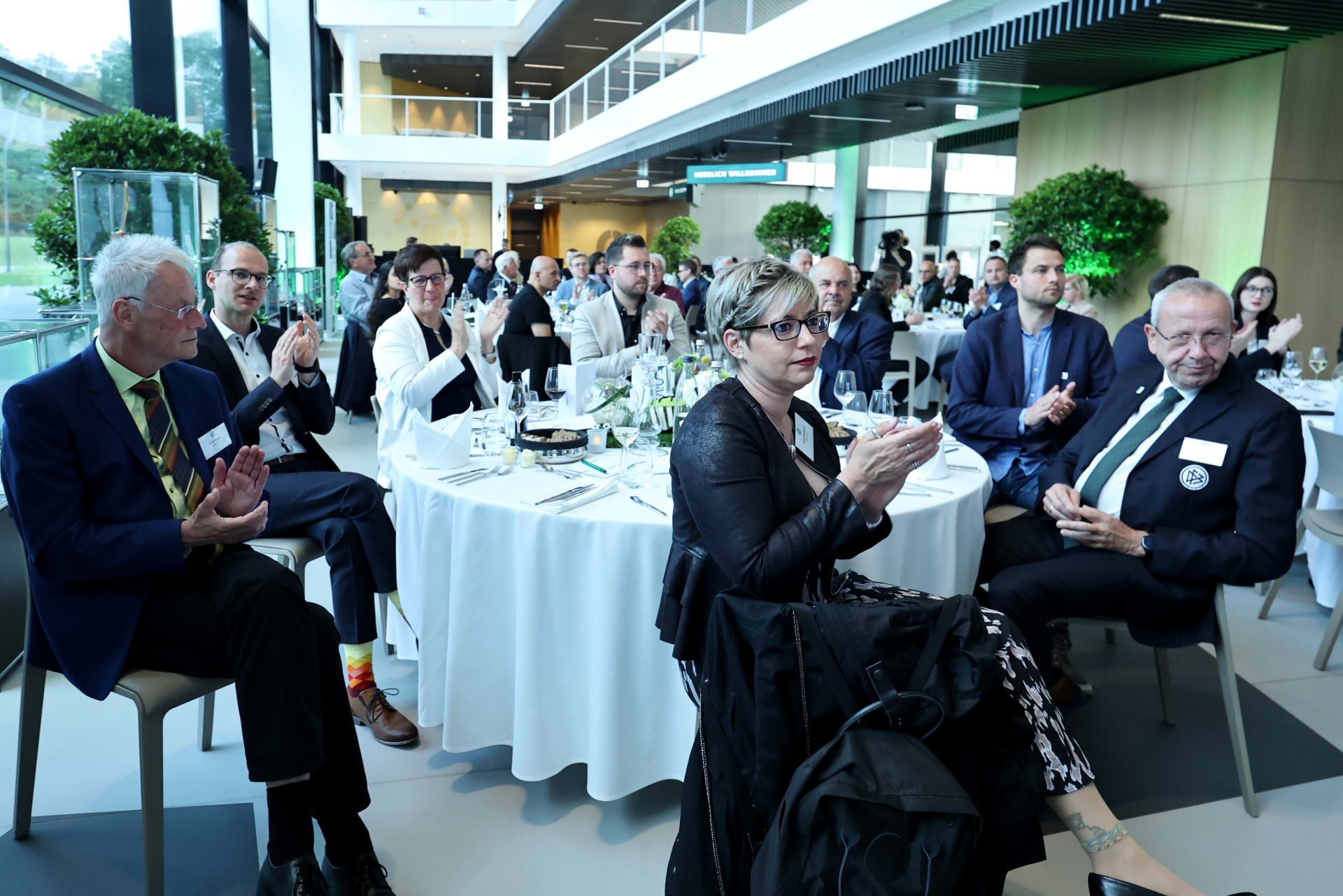 [{"clang_id":1,"value":"Innenaufnahme eines Konferenzraums mit etwa 20 Personen an runden, mit weißen Tischdecken gedeckten Tischen. Die Teilnehmer sitzen und applaudieren."}]