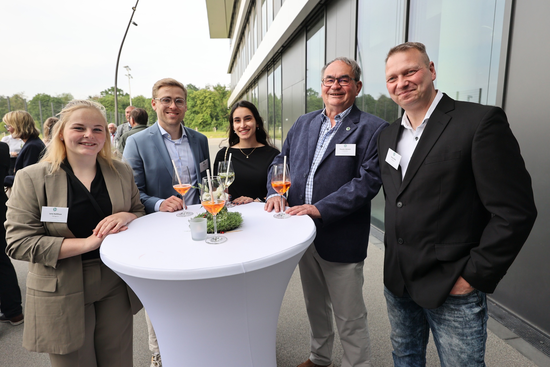 [{"clang_id":1,"value":"Fünf Personen stehen lächelnd vor einem weißen Stehtisch mit Getränken. Im Hintergrund ein modernes Gebäude und Bäume. Alle tragen Businesskleidung mit Namensschildern."}]