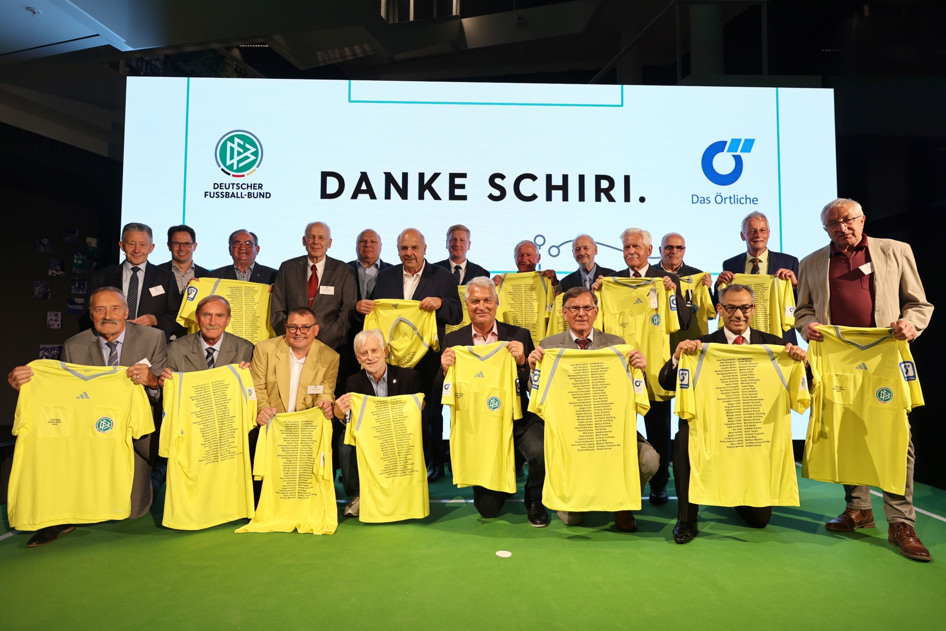 [{"clang_id":1,"value":"Gruppe älterer Männer in Anzügen hält gelbe Trikots mit Text vor einem Banner mit Schriftzug \"Danke Schiri\". Hintergrund ist eine helle Wand."}]