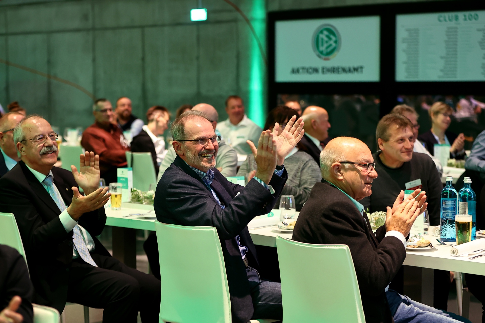 [{"clang_id":1,"value":"Versammlung von Männern in Anzügen, die an Tischen sitzen und klatschen. Im Hintergrund eine grüne Wand mit Logo und Text \"Club 100\"."}]