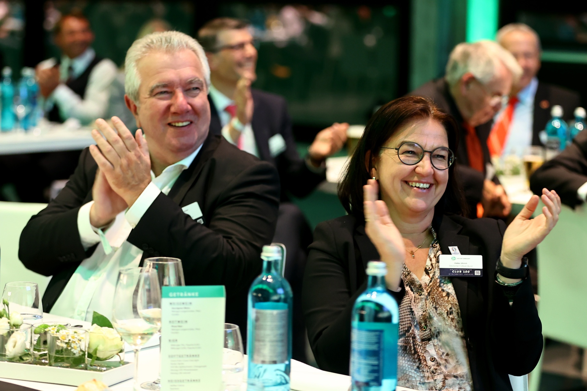[{"clang_id":1,"value":"Zwei Personen applaudieren an einem Tisch in einem Konferenzraum. Im Hintergrund weitere Personen und Tische."}]