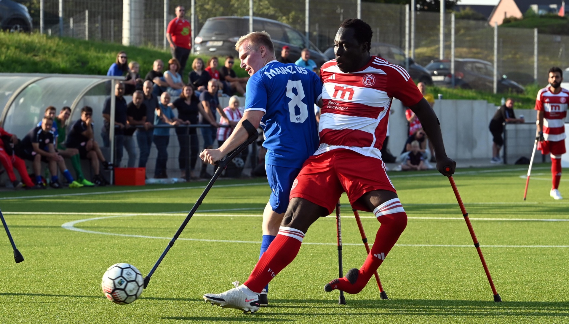 [{"clang_id":1,"value":"Zwei Fußballspieler mit Krücken kämpfen um den Ball auf einem grünen Rasenplatz. Im Hintergrund Zuschauer und ein Stadiondach."}]