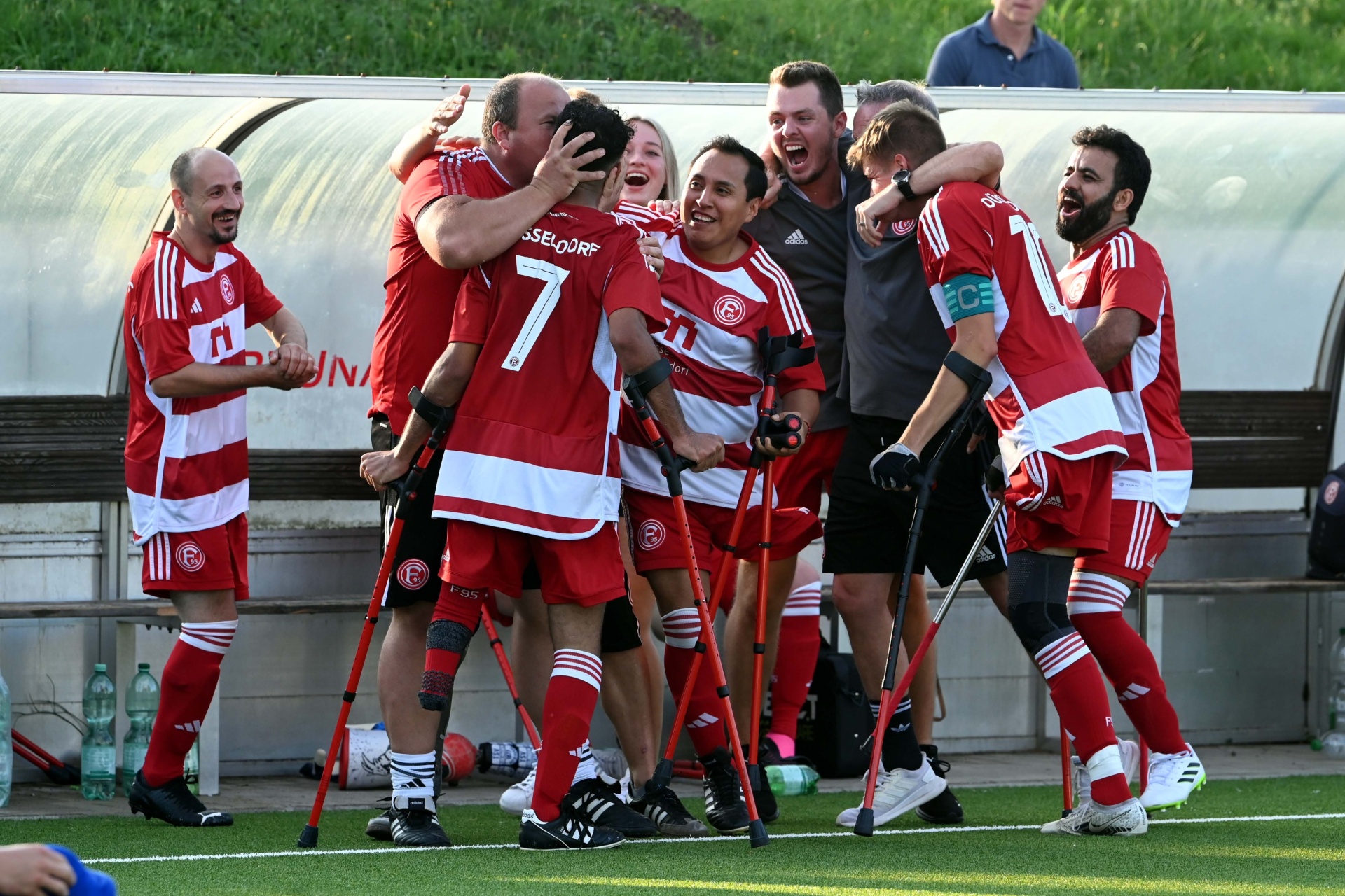 [{"clang_id":1,"value":"Fußballteam in roten Trikots feiert auf dem Spielfeld. Eine Frau mit Krücken wird in die Luft gehoben. Im Hintergrund eine Trainerbank und Zuschauer."}]