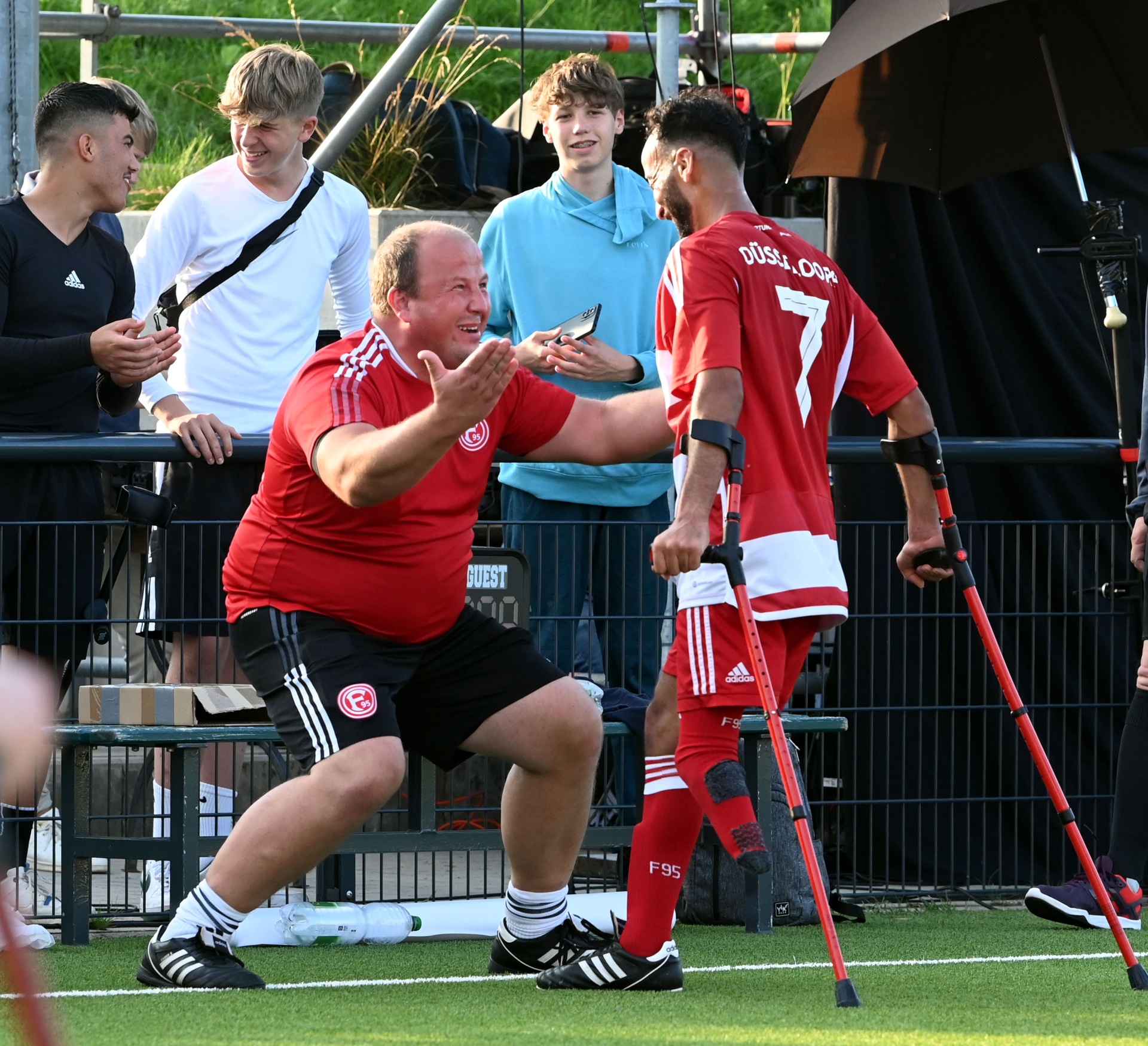 [{"clang_id":1,"value":"Zwei Männer in roten Fußballtrikots. Einer stützt sich auf Krücken, der andere jubelt ihm zu. Im Hintergrund weitere Personen und ein Zaun."}]