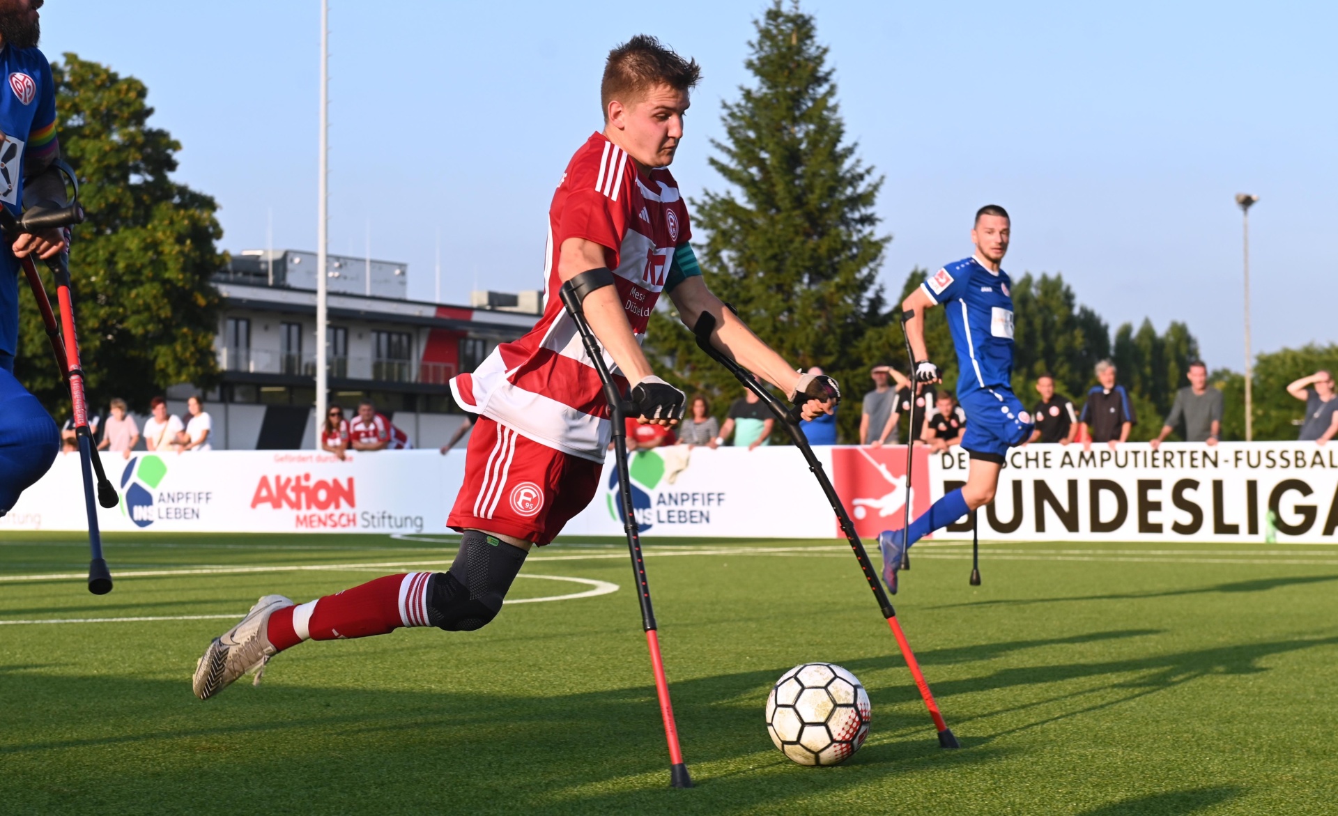 [{"clang_id":1,"value":"Fußballspieler mit Prothesen auf dem Rasen. Er dribbelt den Ball, trägt rot-weißes Trikot und Schienen. Im Hintergrund weitere Spieler und Zuschauerränge."}]