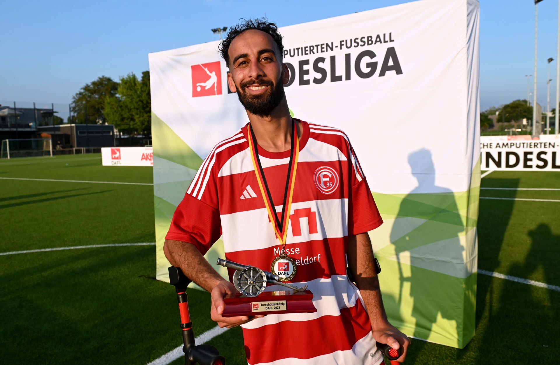 [{"clang_id":1,"value":"Mann mit Goldmedaille und Pokal auf Fußballfeld. Er trägt rot-weiß gestreiftes Trikot und ist lächelnd abgebildet. Im Hintergrund eine Werbeplane und das Spielfeld."}]