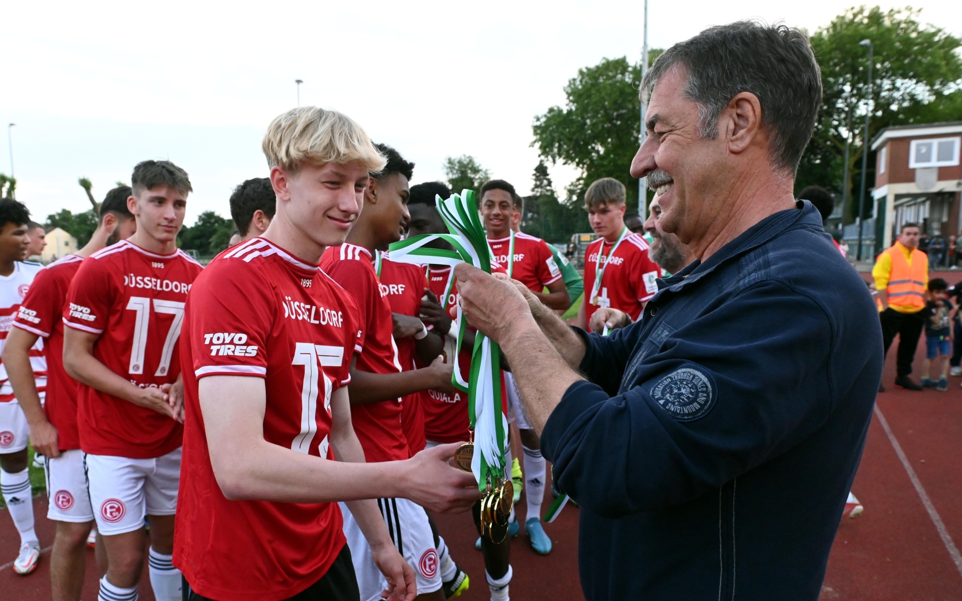 [{"clang_id":1,"value":"Fußballteam in roten Trikots nimmt Medaillen und Urkunden von einem Mann entgegen. Im Hintergrund Zuschauer und Bäume."}]