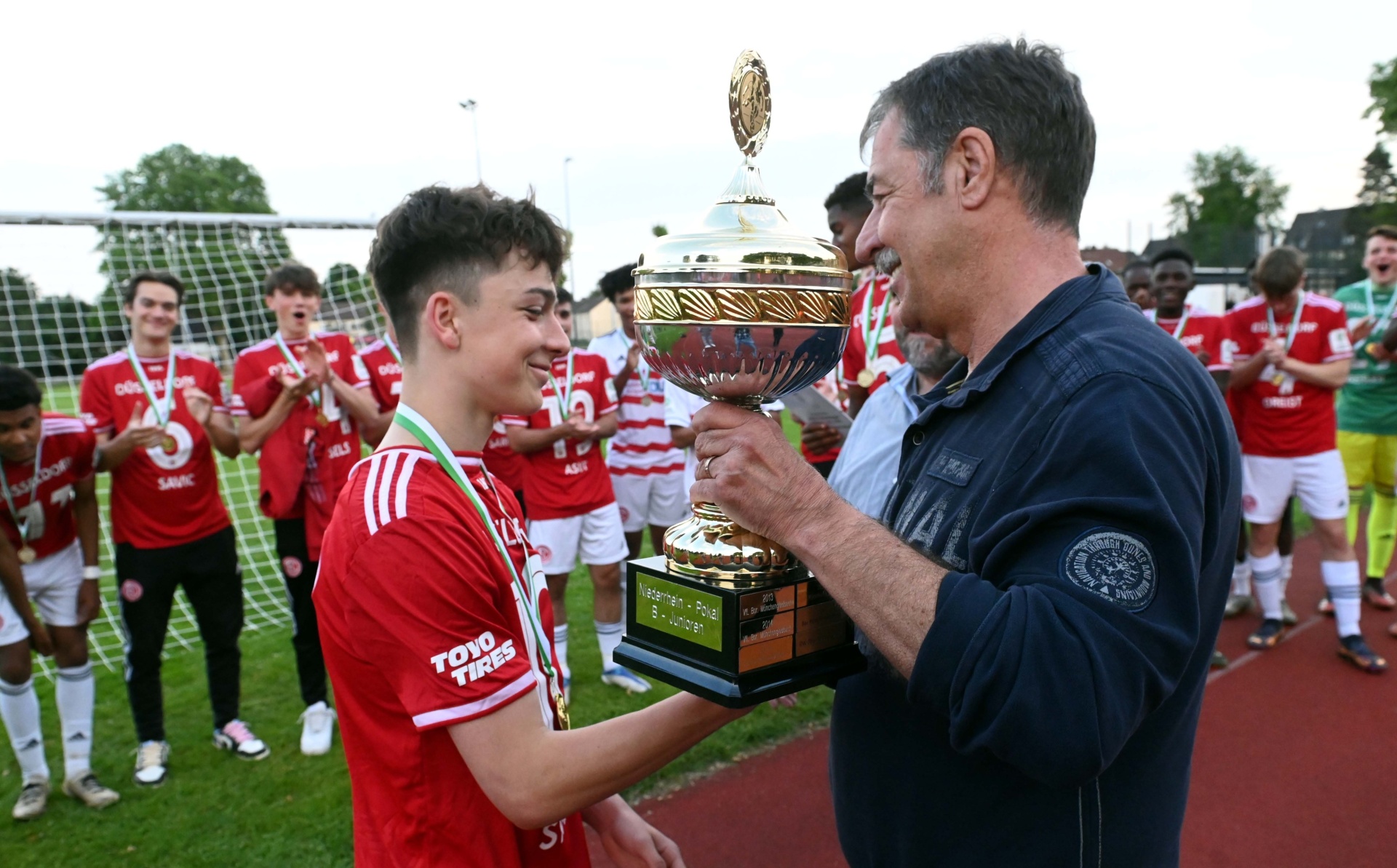 [{"clang_id":1,"value":"Junge im Fußballtrikot nimmt Pokal von Mann entgegen. Im Hintergrund stehen jubelnde Fußballspieler auf einem Rasenfeld."}]