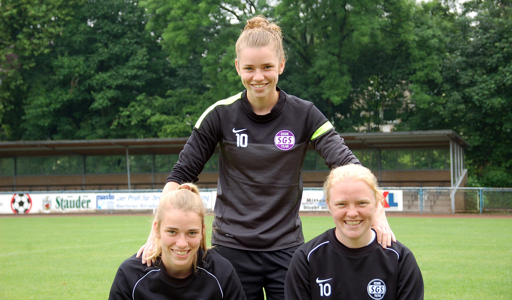 [{"clang_id":1,"value":"Drei Frauen in schwarzen Sportanzügen mit pinkem Logo stehen auf einem Fußballfeld vor Bäumen. Die mittlere Frau sitzt, die anderen stehen dahinter."}]