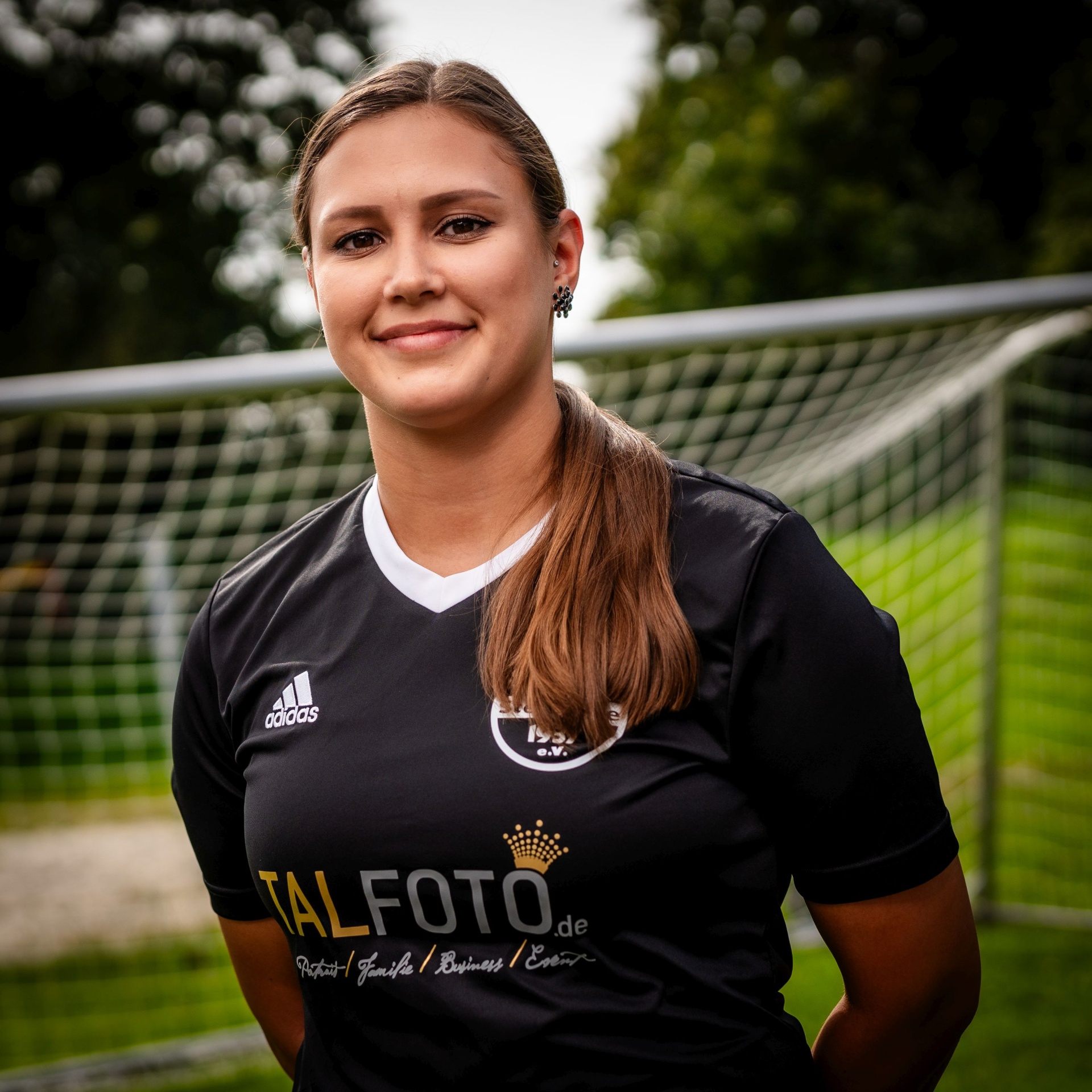 [{"clang_id":1,"value":"Frau in schwarzem Fußballtrikot mit weißem Kragen und Ärmeln lächelt in die Kamera. Im Hintergrund ein Fußballtor und grüne Vegetation."}]