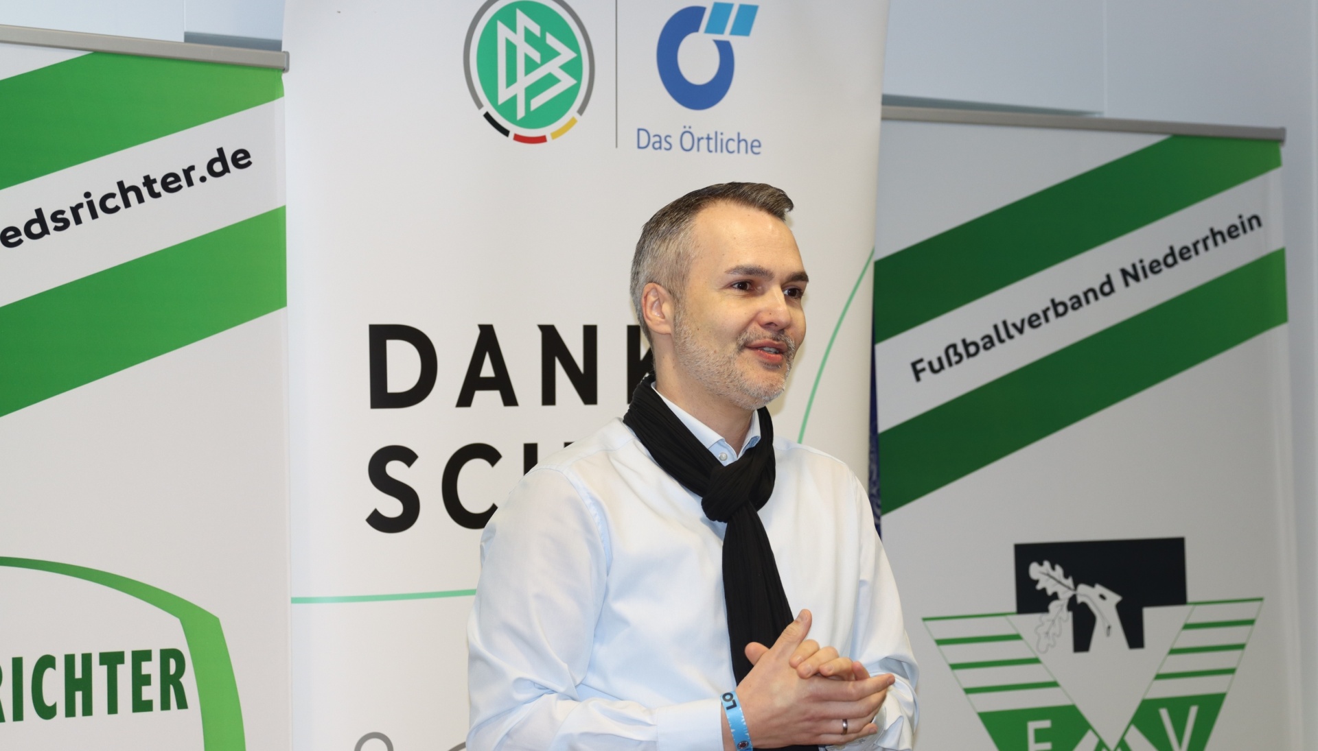 [{"clang_id":1,"value":"Mann mit schwarzem Schal und weißem Hemd vor Werbebannern mit Fußball-Logos und Texten. Im Hintergrund grüne Banner mit Schriftzügen."}]